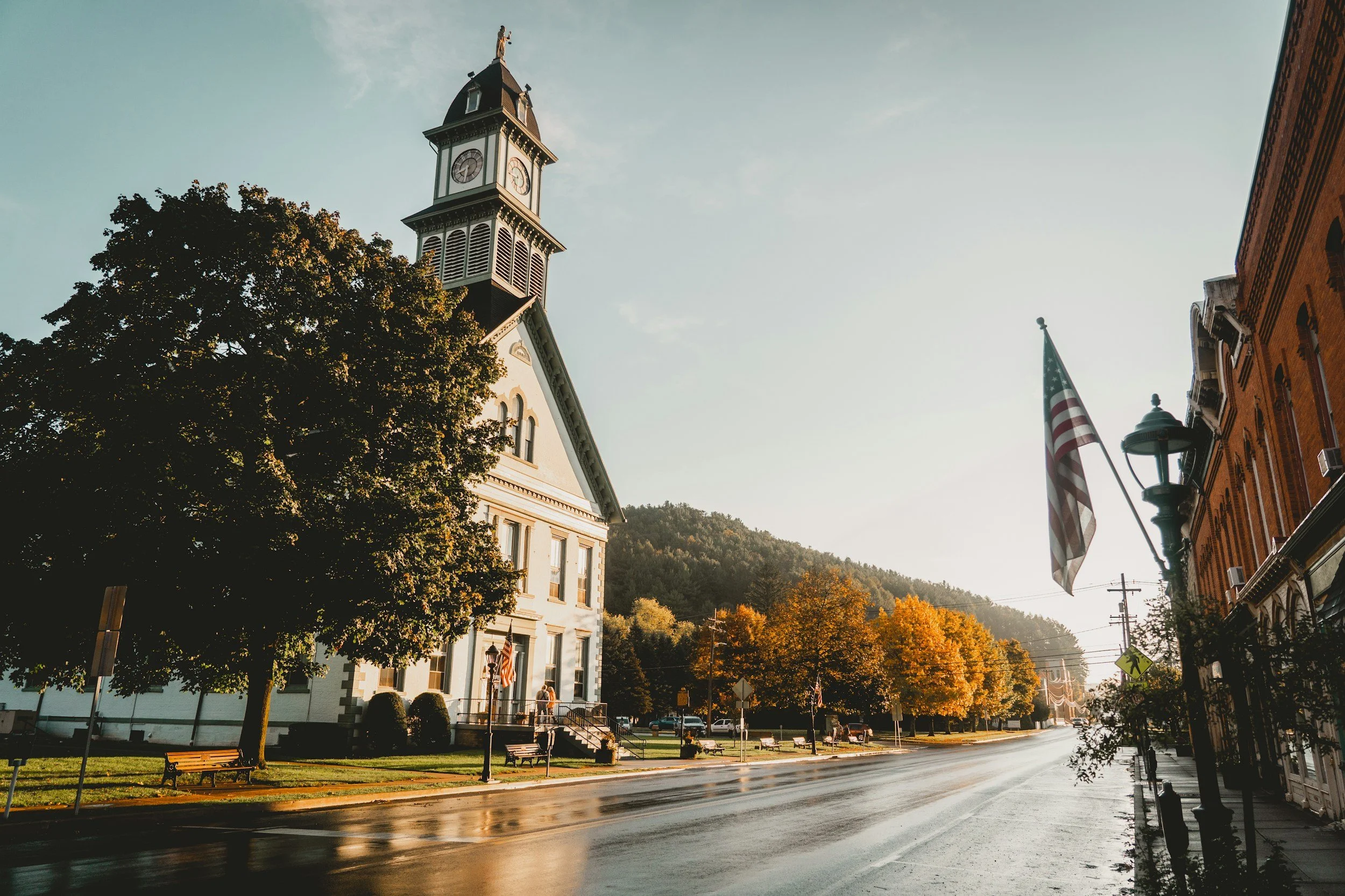 American small town with a historic clock tower church, American flag, brick buildings, benches, and autumn trees with colorful leaves