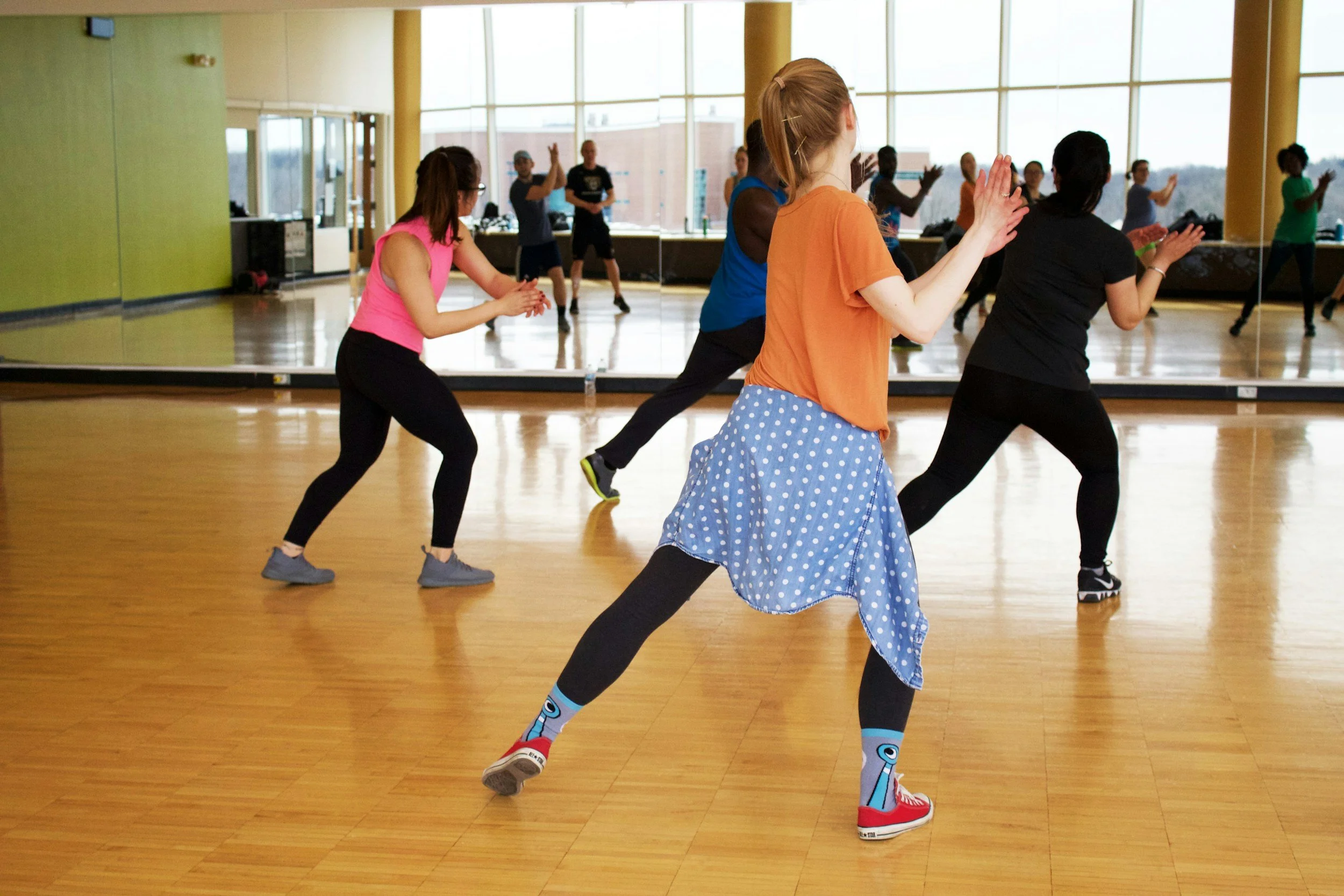 People participating in a dance or exercise class in a fitness studio with large windows and wooden floors.