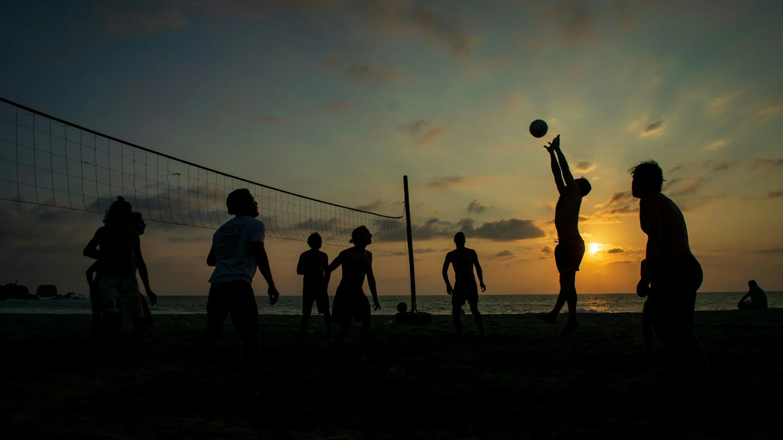 Silhouettes of people playing beach volleyball at sunset with the ocean in the background.