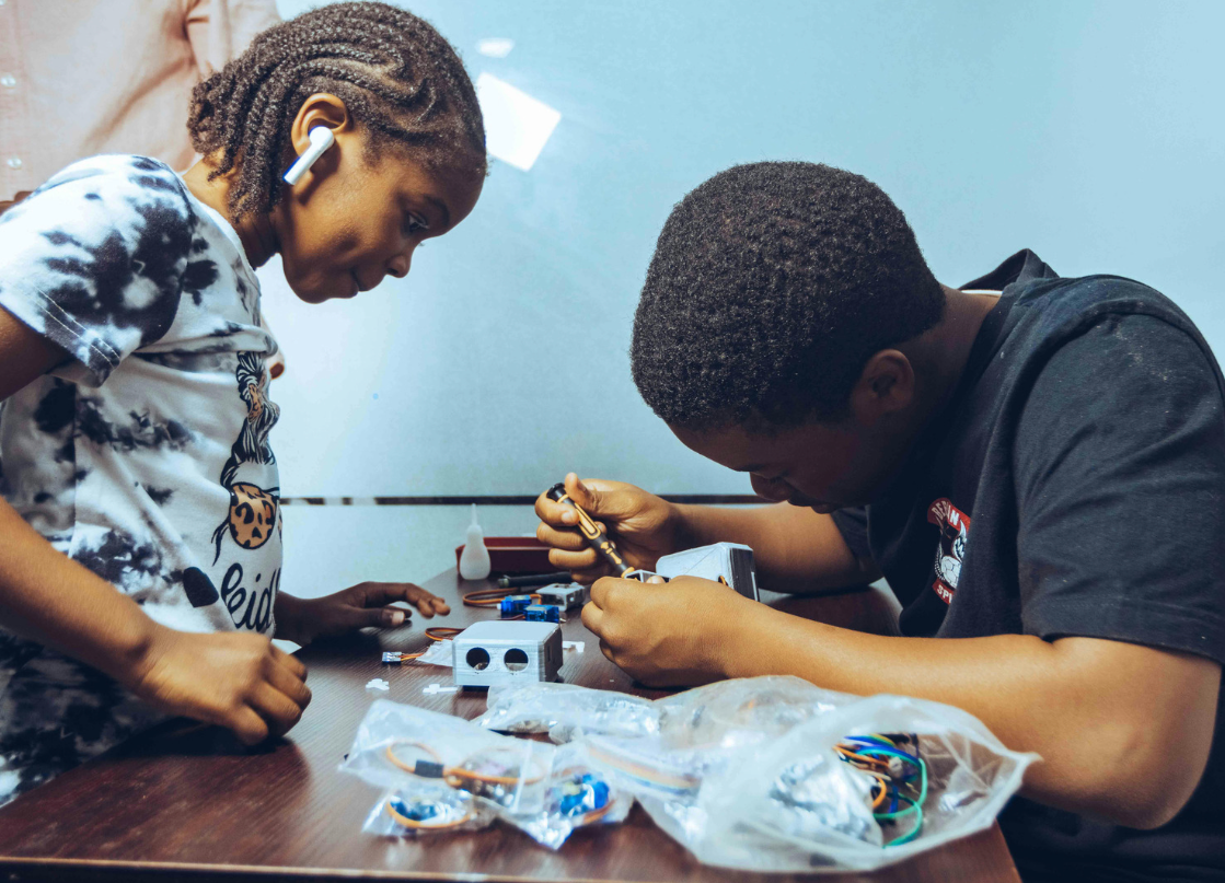 Two individuals working on an electronic project at a table with various tools and electronic parts.