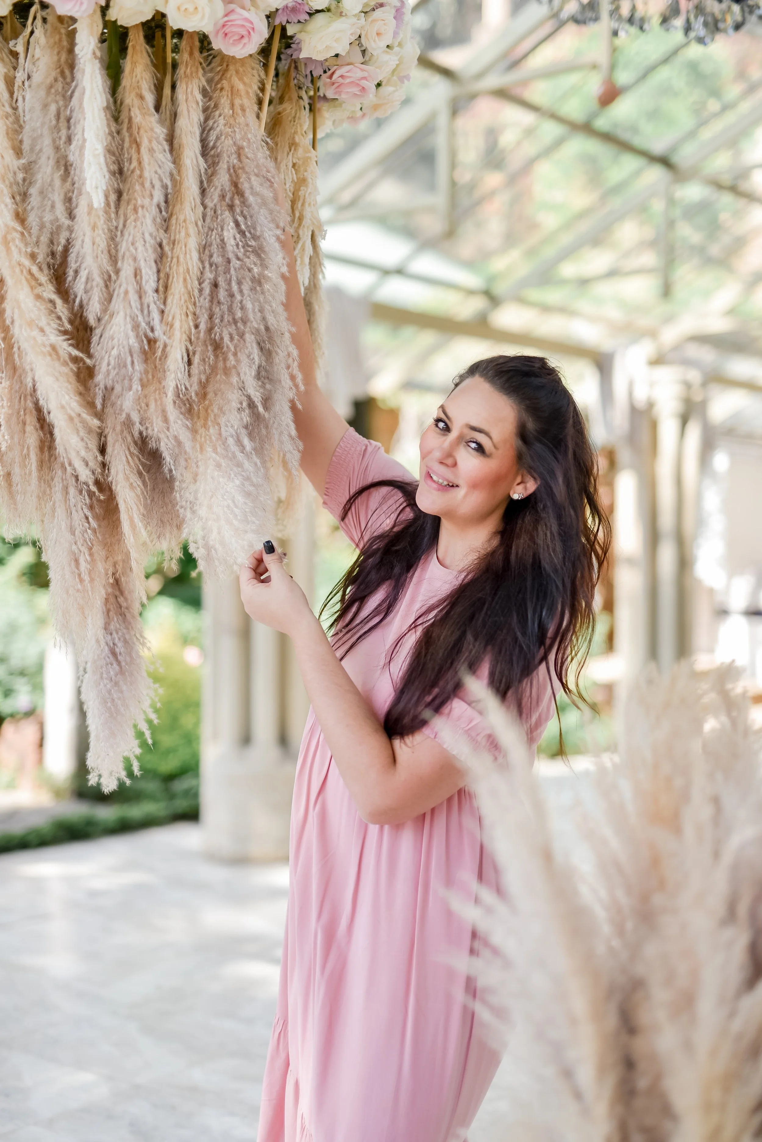 Mira in a pink dress smiling and touching hanging floral and pampas design, in a bright, indoor setting with a glass ceiling and classical columns.