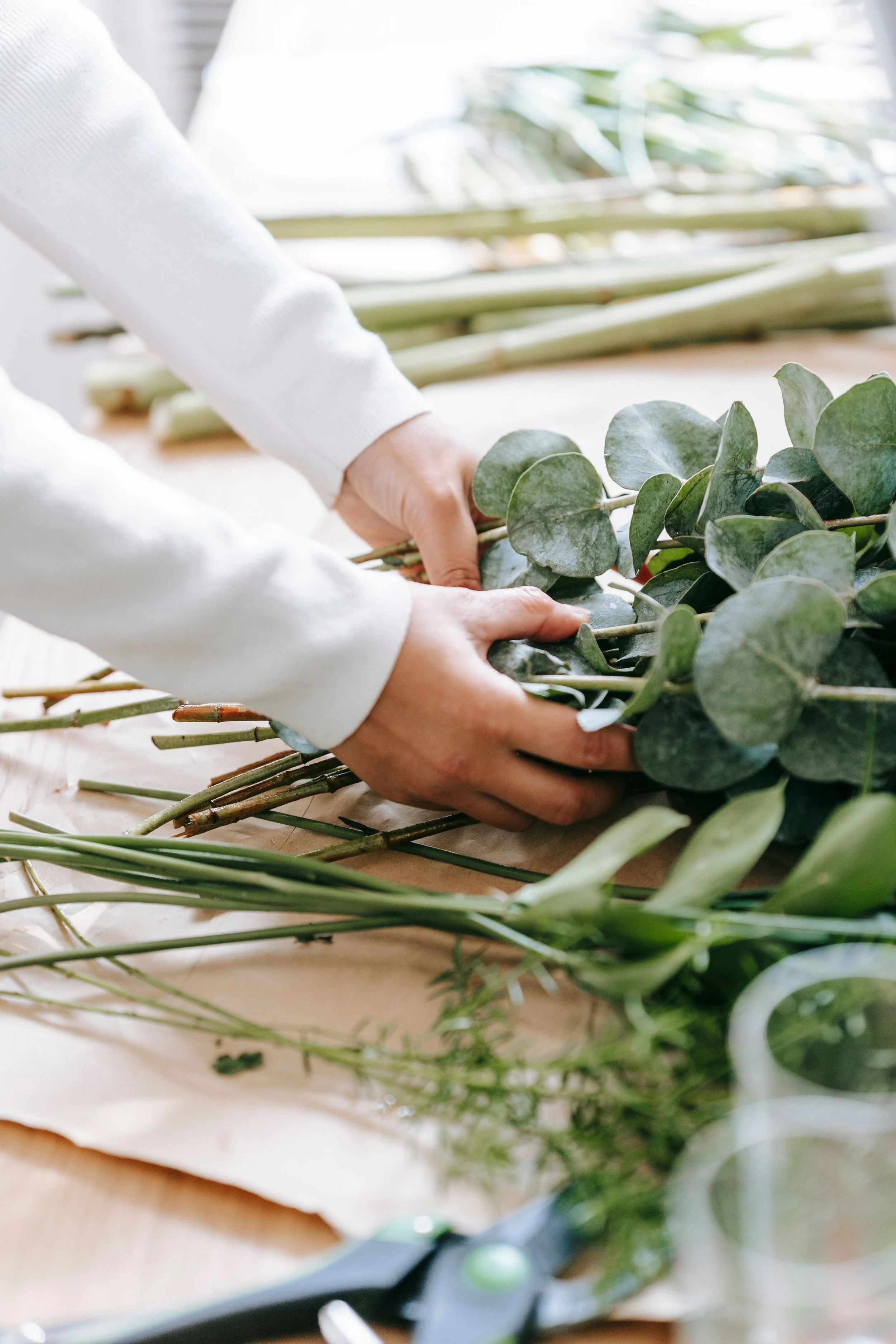 Person arranging eucalyptus and green leafy stems on a table for floral arrangement or decoration.