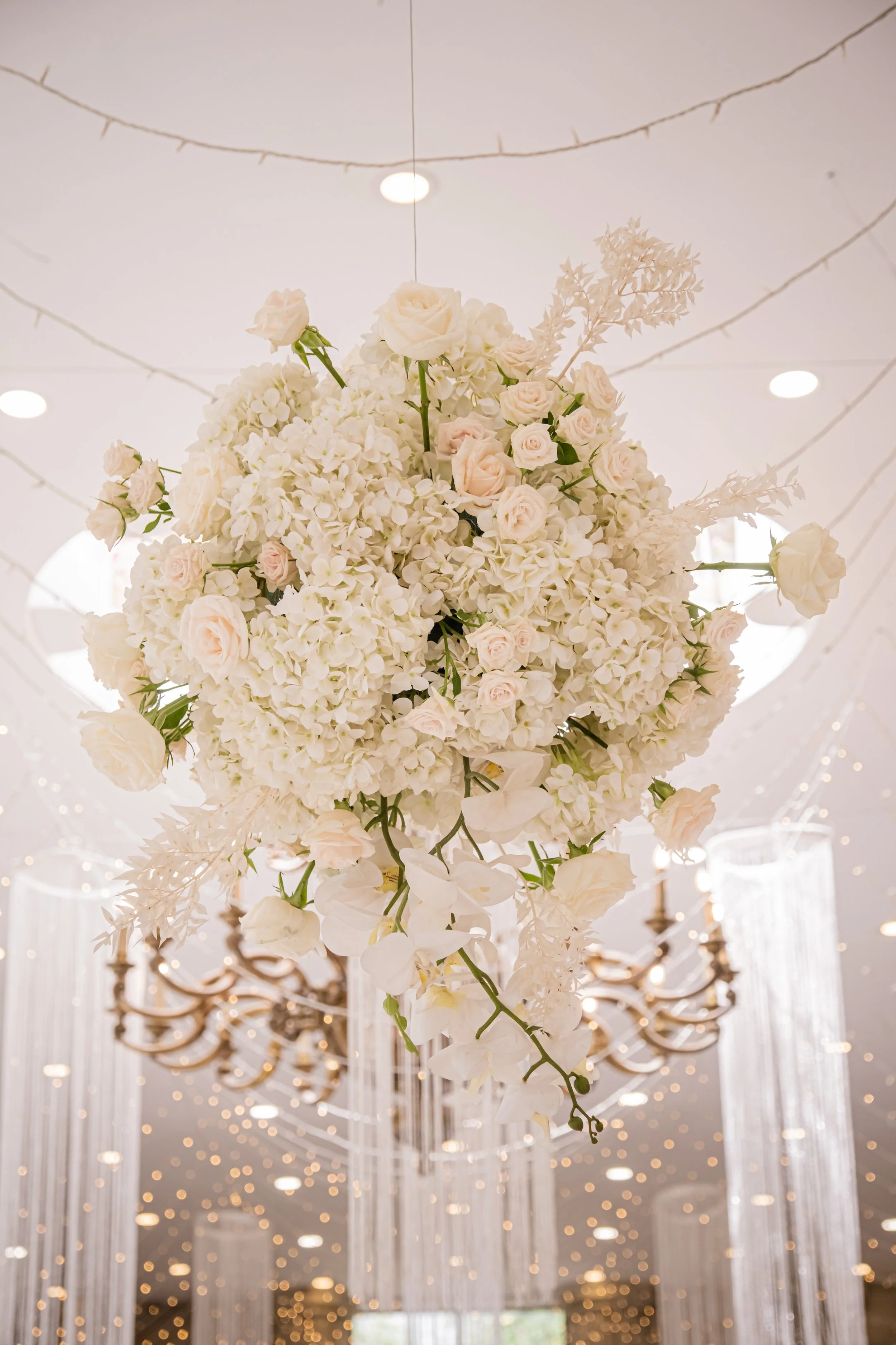A large floral centerpiece of white and light pink roses and hydrangeas hanging from a ceiling decorated with string lights, with chandeliers and draped curtains in the background.