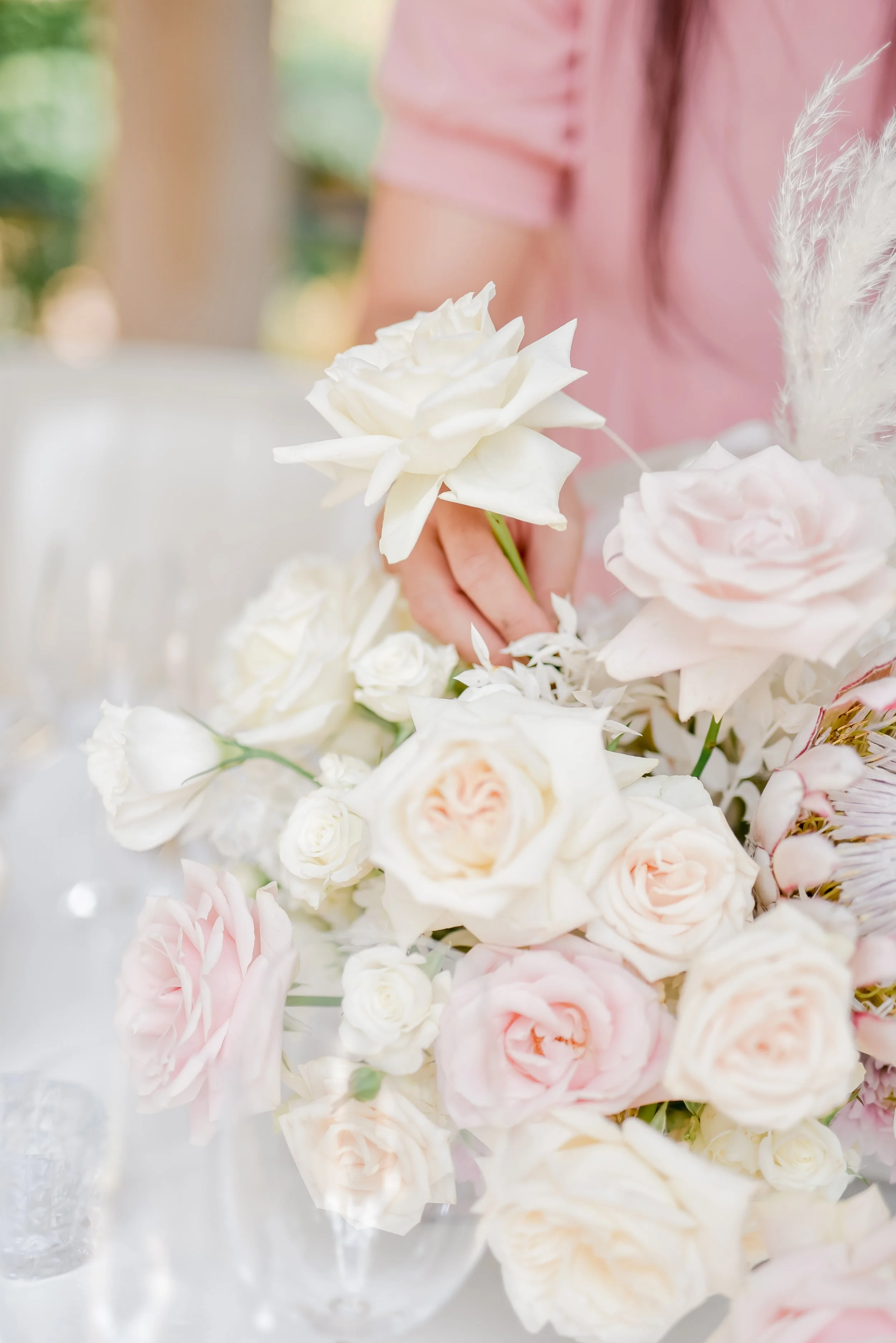 Mira in a pink dress arranging a bouquet of white and light pink roses and other flowers at a table.