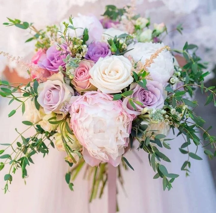 Close-up of a pastel flower bouquet with pink, white, and lavender roses, peonies, and greenery against a soft background.
