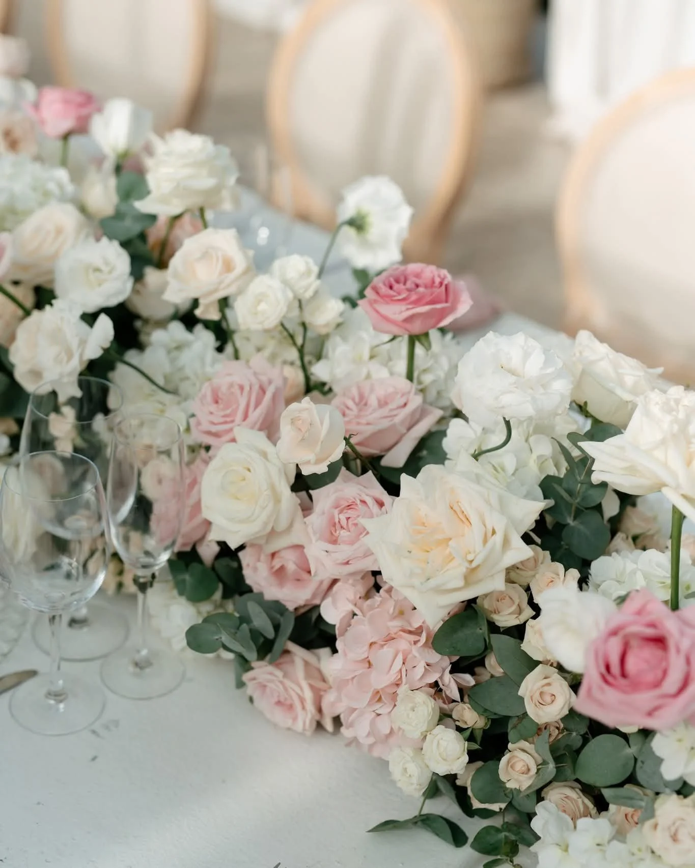Close-up of a floral centerpiece with pink, white, and cream roses and greenery on a table, with empty wine glasses nearby, in a softly lit setting.