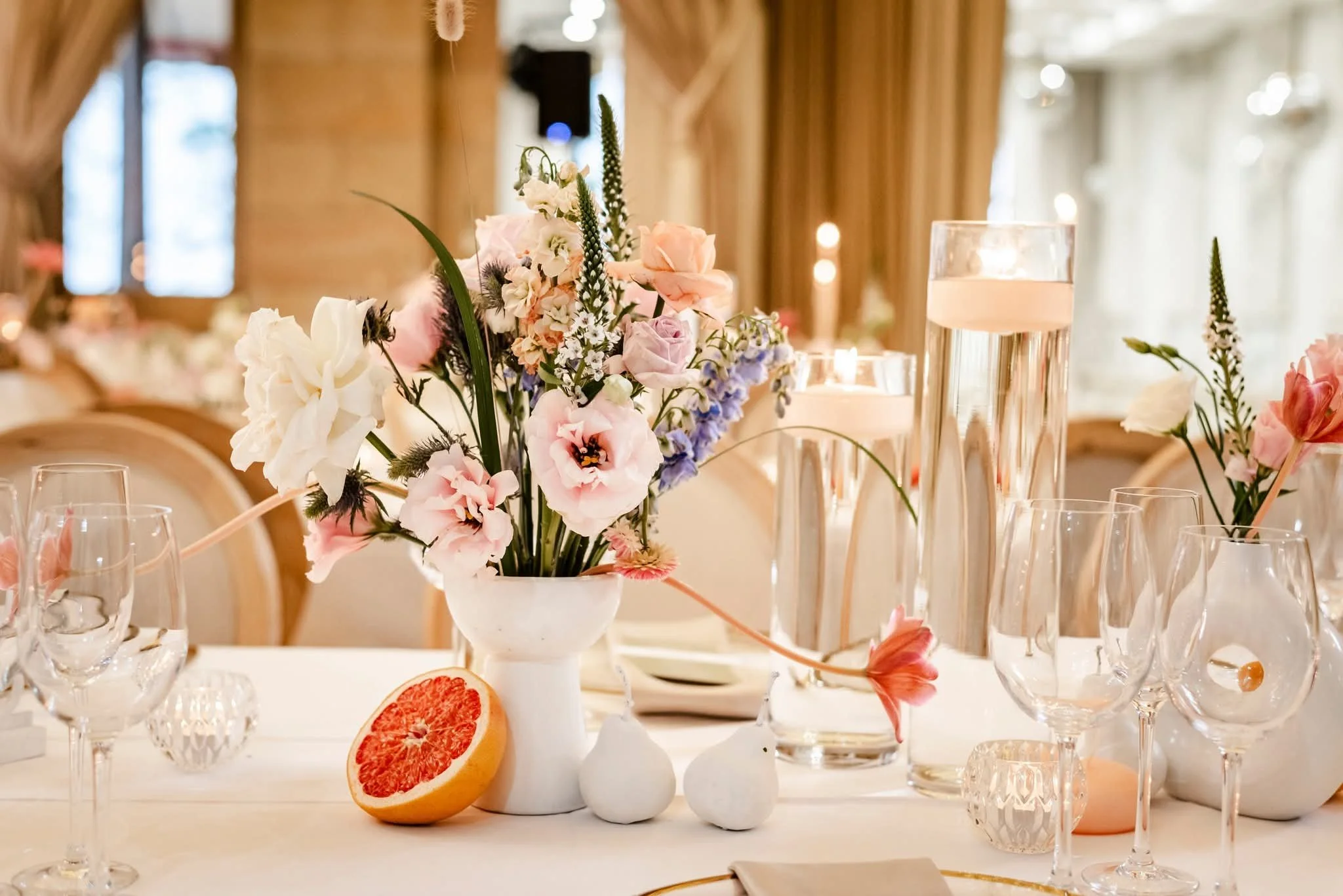 Elegant table centerpiece featuring a bouquet of pink, white, and purple flowers in a white vase, surrounded by clear glassware, with a halved grapefruit in the foreground and tall glass candleholders, set in a warmly lit room with wooden architectural details.