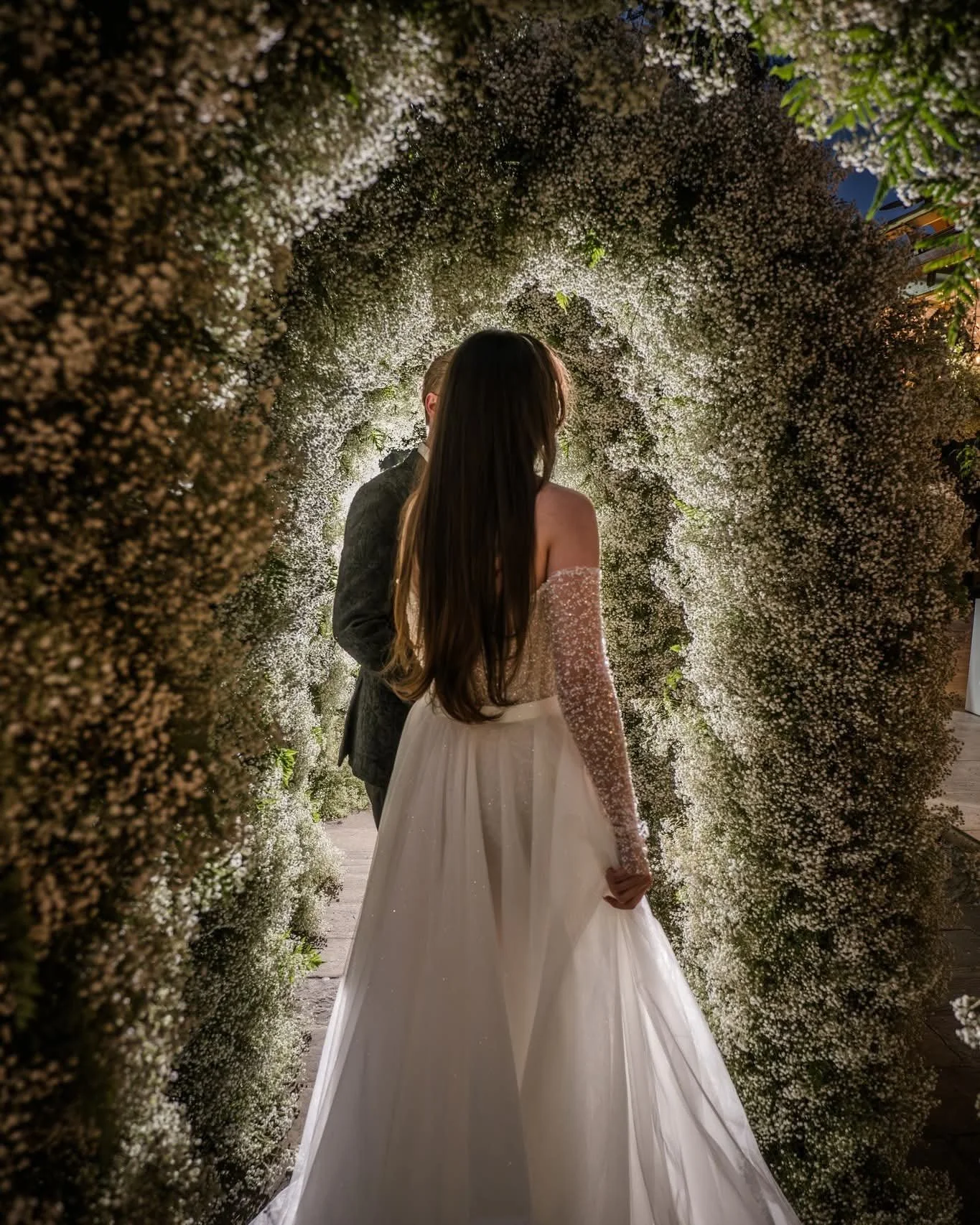 A bride and groom holding hands under an archway made of white flowers during a wedding ceremony at night.