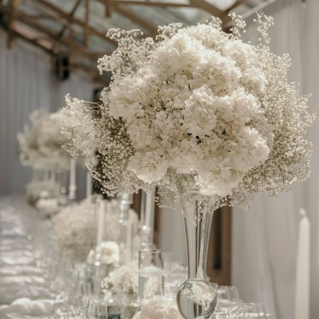 A large elegant bouquet of white flowers, including hydrangeas and baby's breath, arranged in a tall, clear glass vase on a decorated table for a formal event.