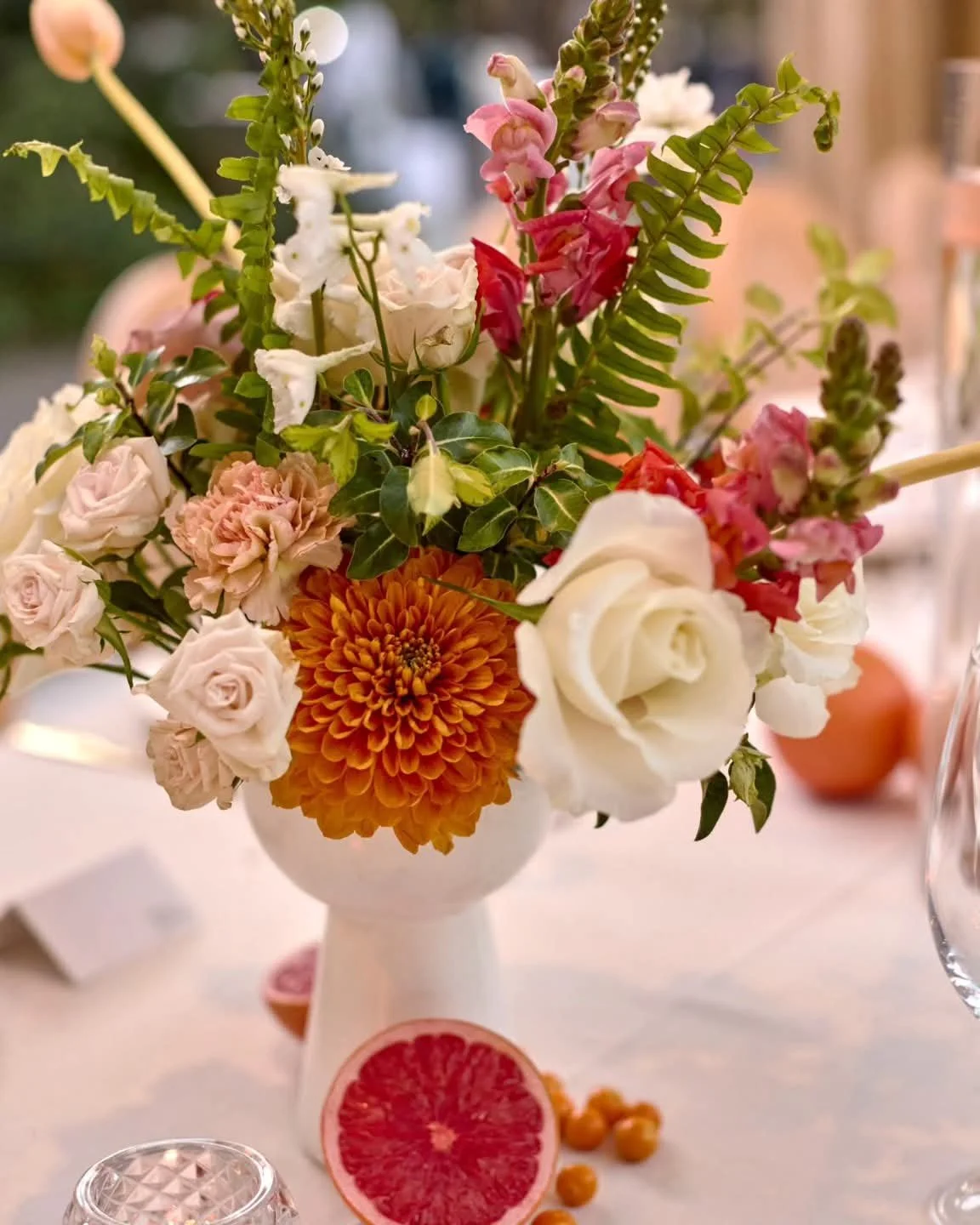 A floral bouquet in a white vase with pink, white, and orange flowers, including roses, a chrysanthemum, and other blooms, placed on a table with sliced grapefruit and scattered small orange berries.