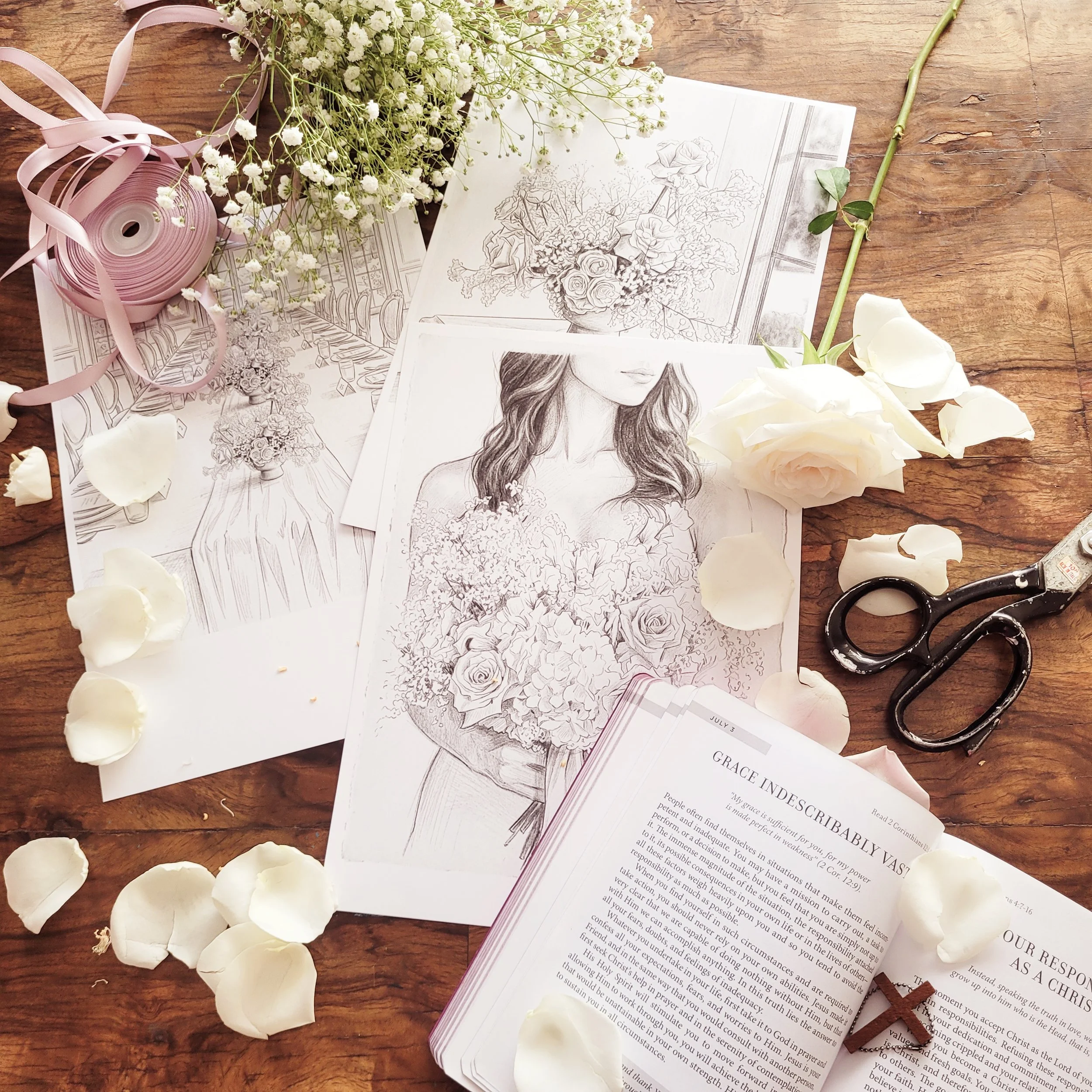 Study or work space with sketches of floral arrangements, wedding dress, and portrait of a woman, surrounded by white roses, baby's breath flowers, scissors, a pink ribbon spool, and a rosary on a wooden surface.