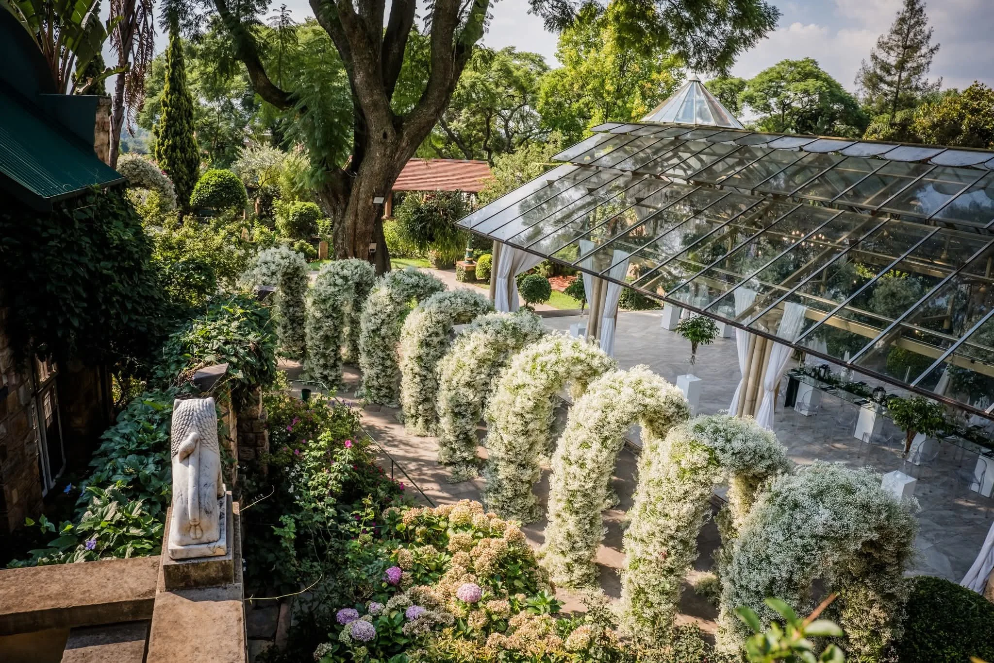 A lush garden with a large tree, various bushes, and flowering plants, featuring a glass greenhouse structure and chairs underneath it. Alongside the glass structure twelve magnificent created babies breathe arches.