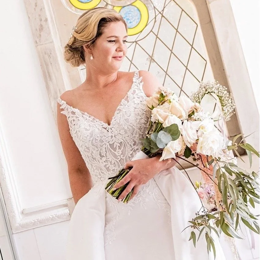 A bride in a white lace wedding gown holding a bouquet of white and blush roses and greenery, standing indoors near a decorated wall with a stained glass window in the background.