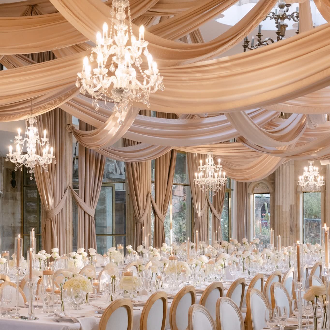 Elegant banquet hall decorated for a wedding or special event with draped fabric ceiling, crystal chandeliers, tall windows with curtains, and a long table set with white flowers, candles, and glassware.