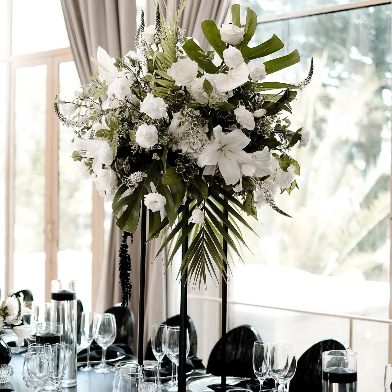 White, and back accented floral centerpiece with greenery on a black stand in a decorated dining area with glasses and chairs, near large windows with curtains.