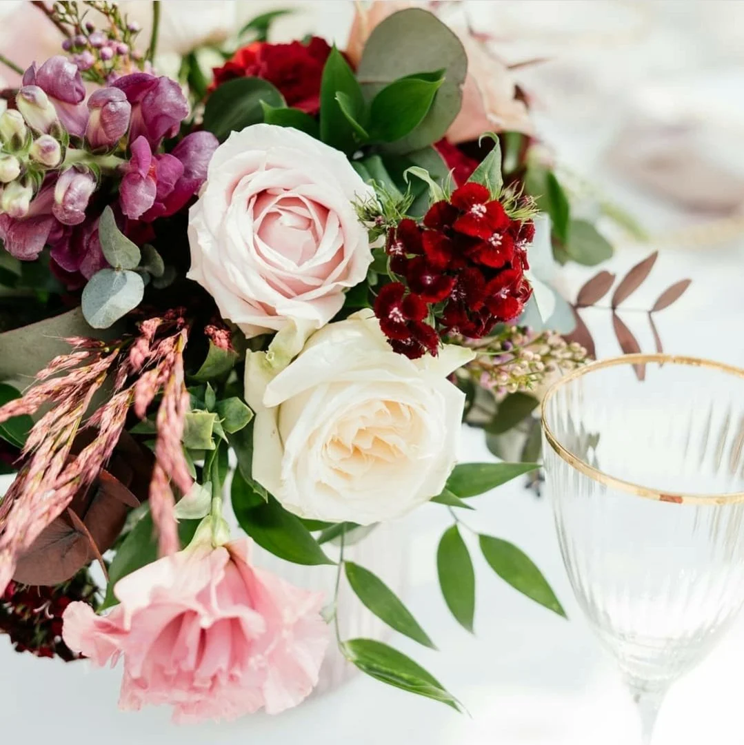 A floral arrangement with pink roses, white roses, red and pink flowers, green leaves, and other plants, next to an empty glass with gold rim on a white surface.