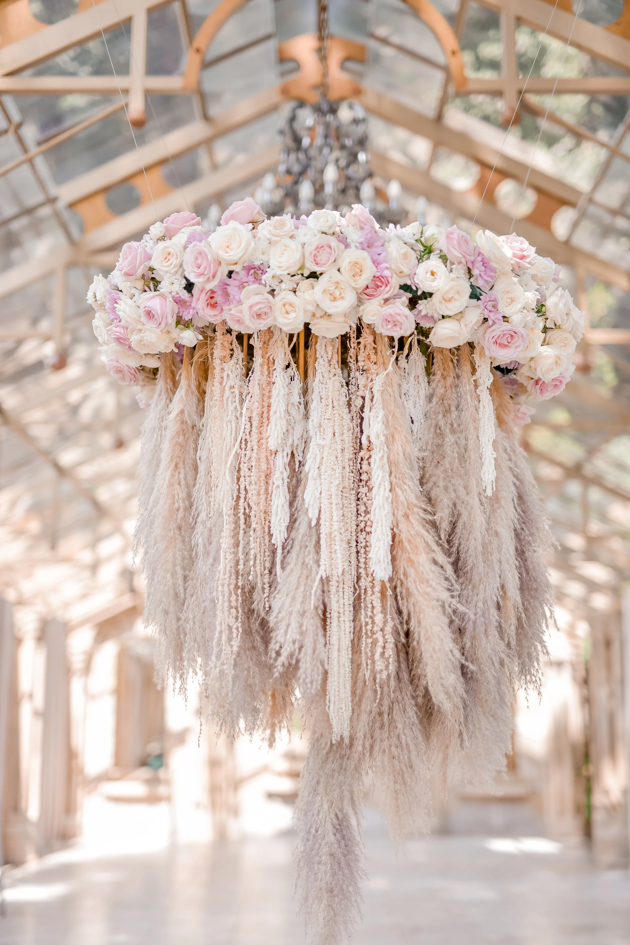 A floral chandelier made of pink and white roses with hanging beige and cream-colored pampas, hanging from the ceiling of a wooden-framed glass-covered structure.