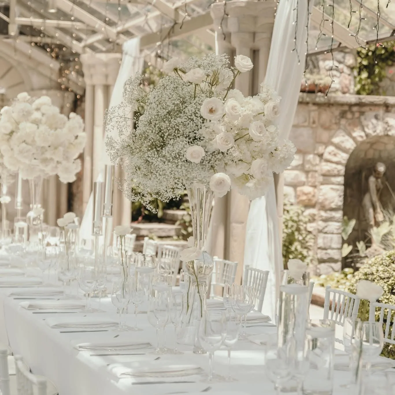 Elegant outdoor wedding reception table with tall floral arrangements of white roses and baby's breath, set with glassware and white chairs, in a venue with stone walls and string lights.