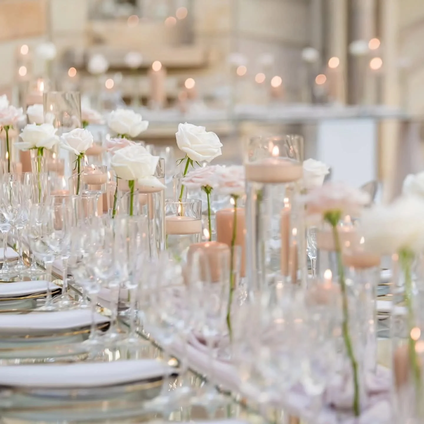 Elegant table setting with white roses in bud vases, candles, and glassware in a softly lit reception area.