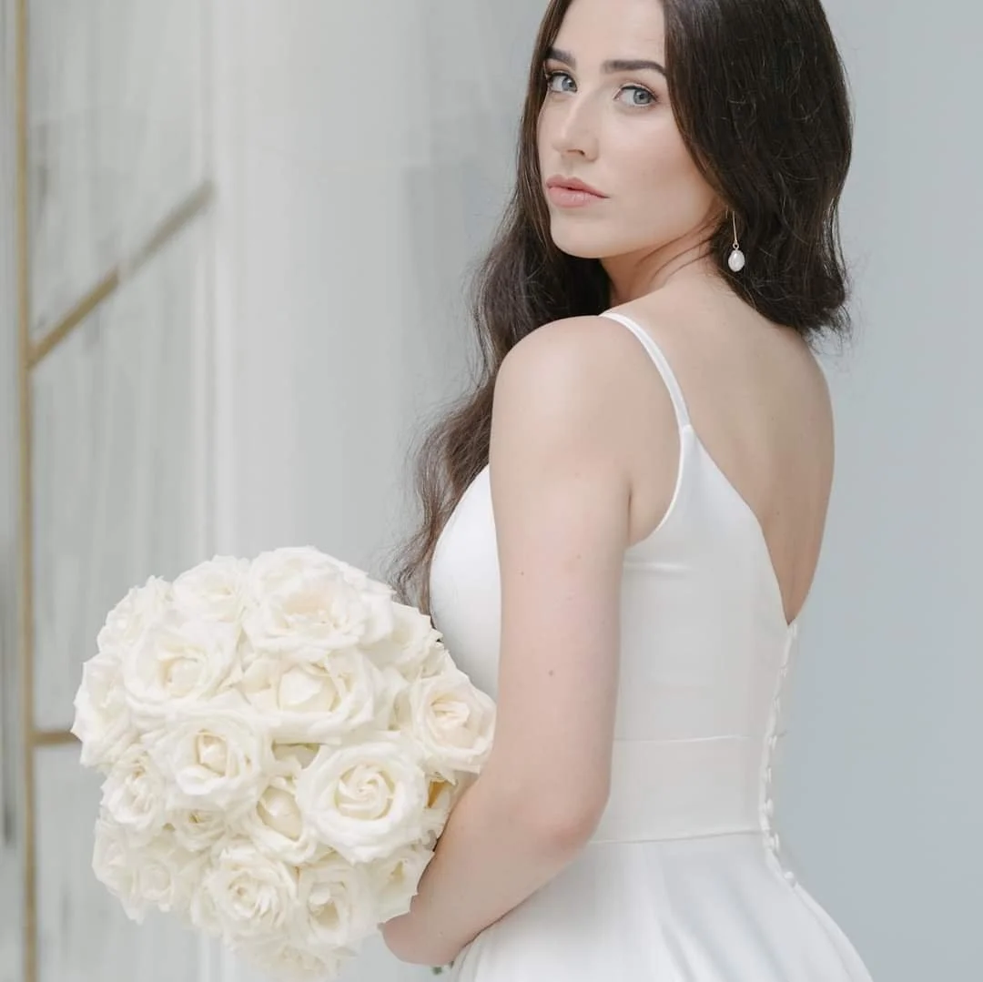 A woman with long dark hair wearing a white wedding dress holding a bouquet of white roses.
