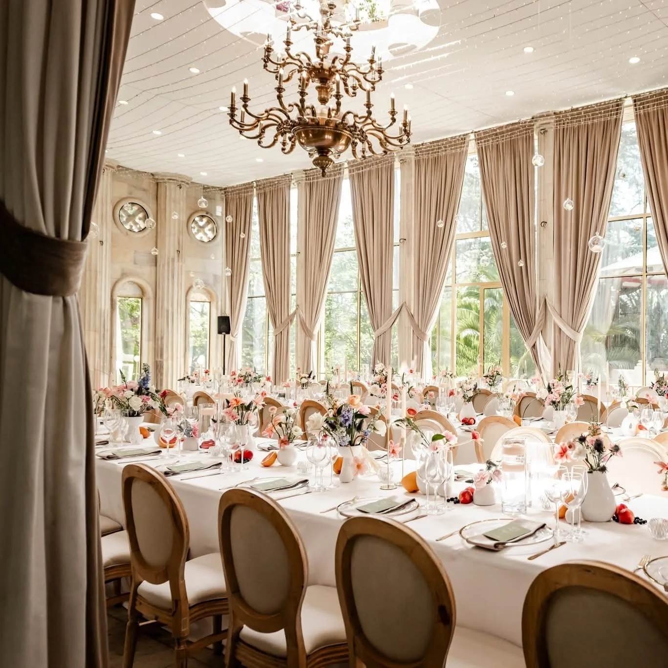 Elegant banquet hall with large windows, long draped curtains, a chandelier hanging from the ceiling, and tables decorated with flowers and place settings.