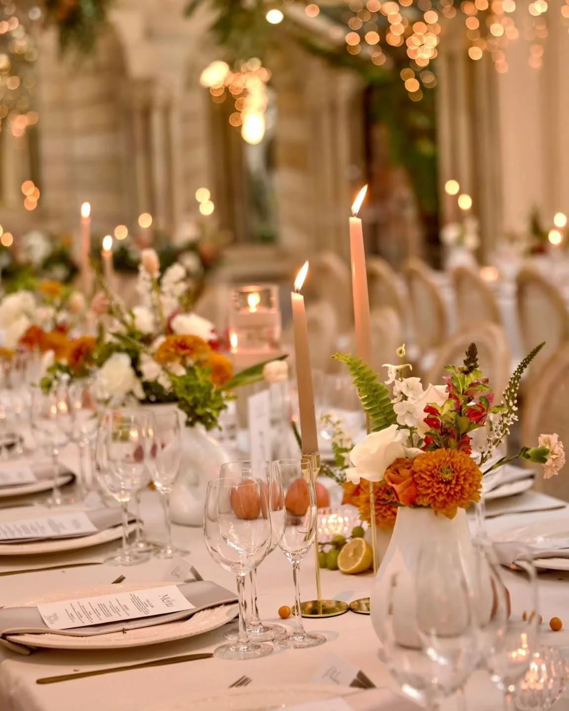 Elegant dining table set with floral arrangements, lit candles, wine glasses, and place settings, in a warmly lit banquet hall.