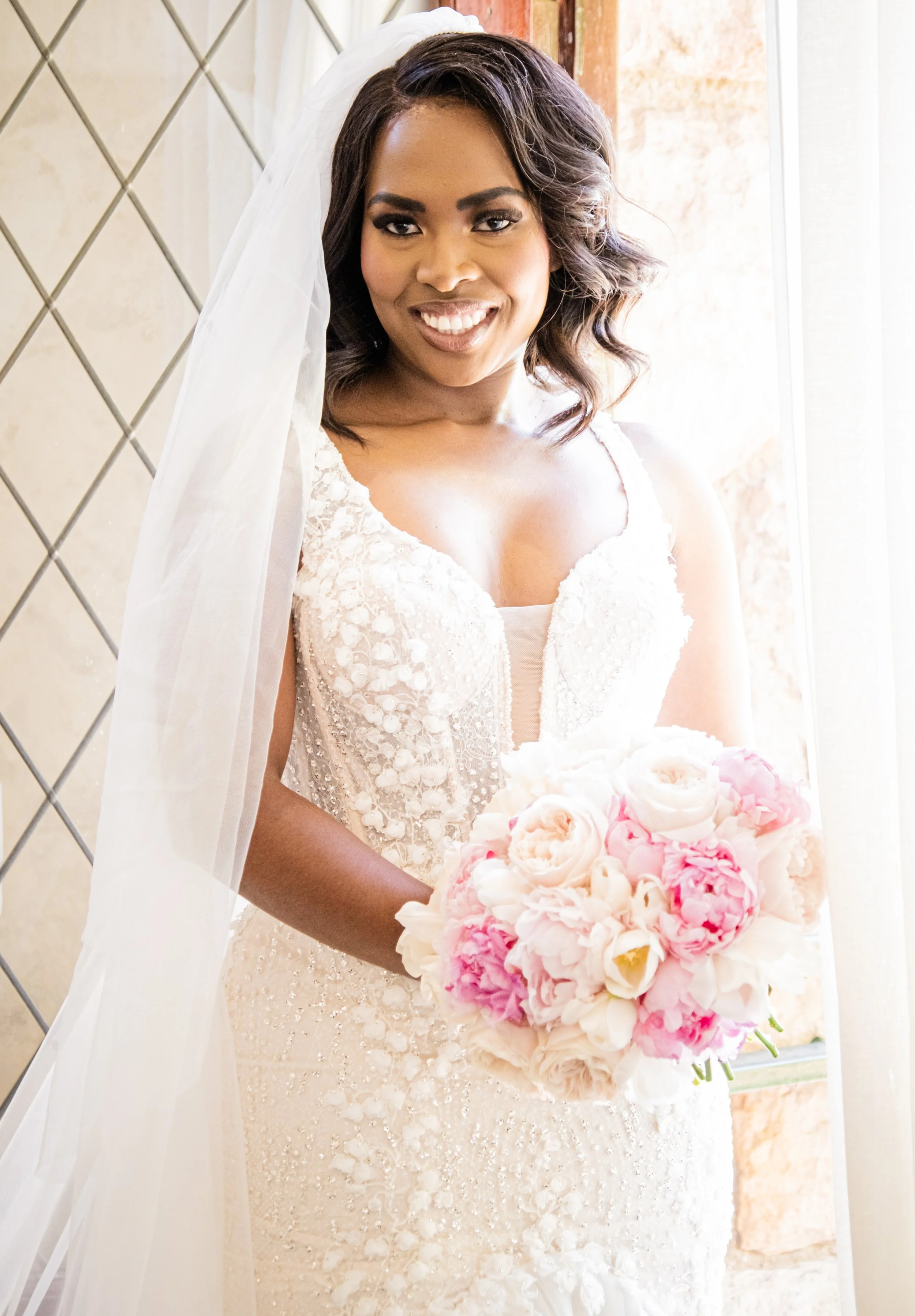 A smiling bride in a white wedding dress holding a bouquet of pink and white peonies.