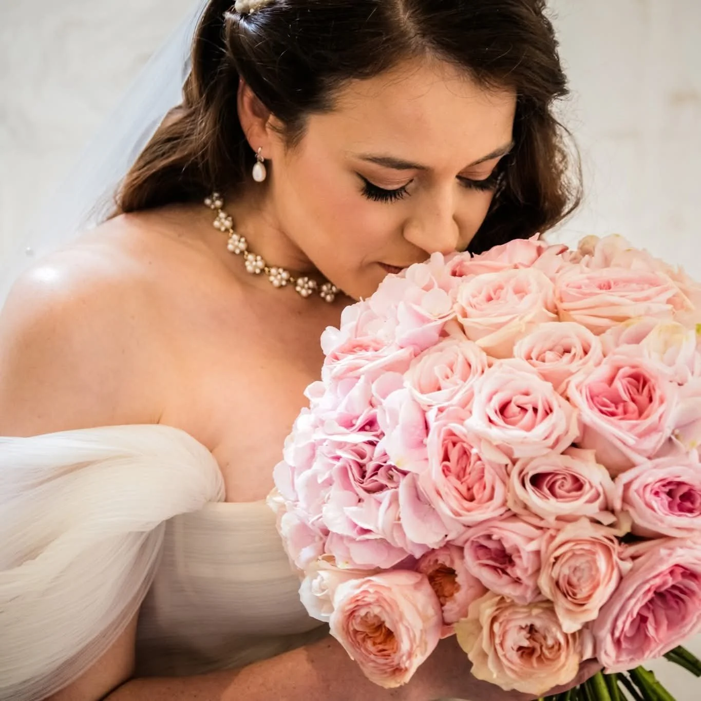 A bride in a white wedding dress holding a large bouquet of pink roses, smelling the flowers with her eyes closed.