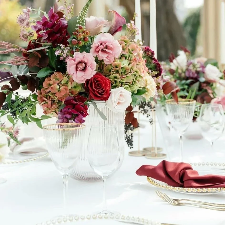 Elegant table setting with pink, white, and red floral centerpiece, empty glassware, gold-rimmed plates, red cloth napkin, and gold-toned cutlery on a white tablecloth.