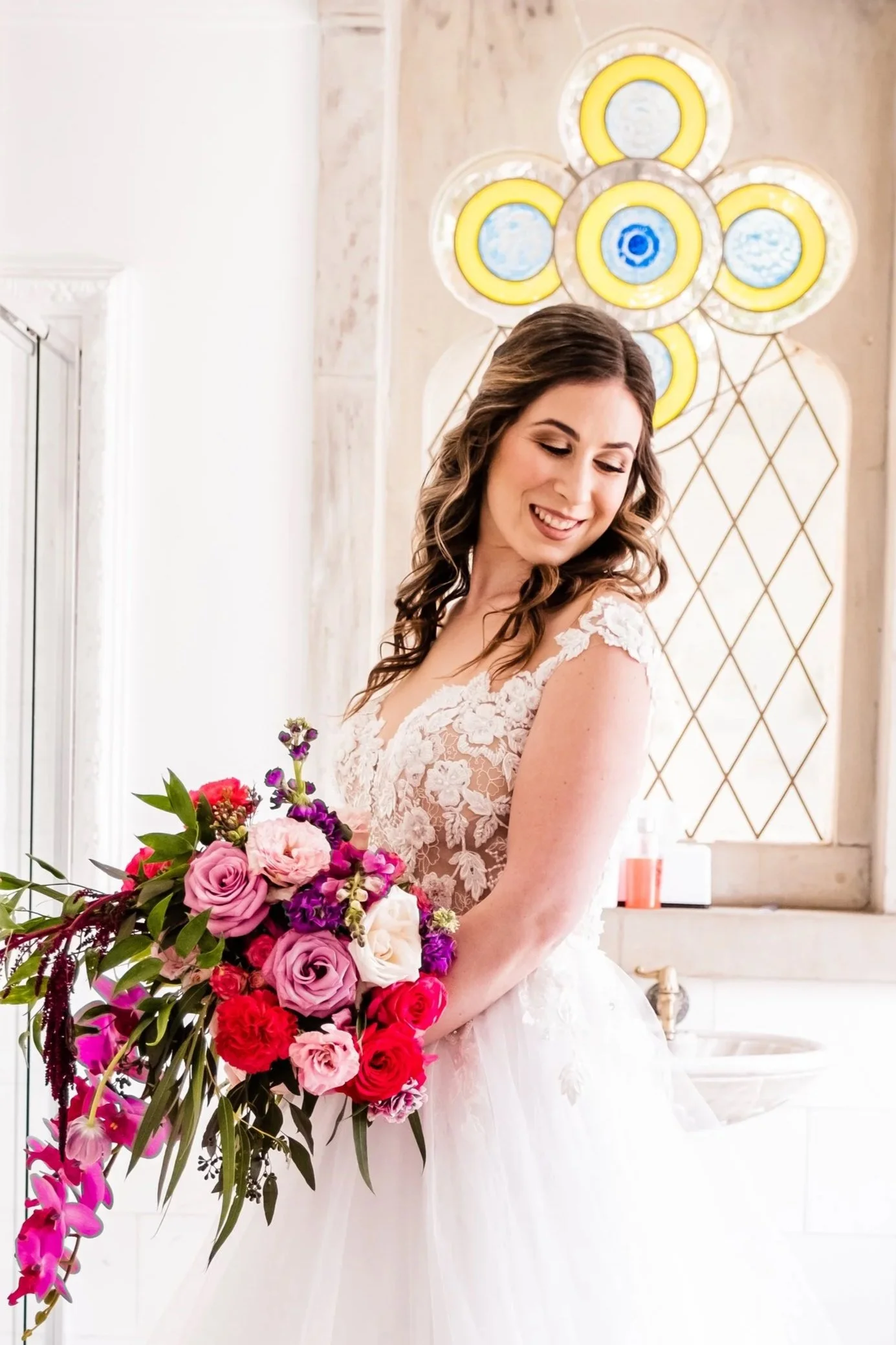 A bride in a white lace wedding dress holding a large bouquet of pink, purple, and white flowers, smiling and looking down, with stained glass window backgrounds and a white marble countertop.