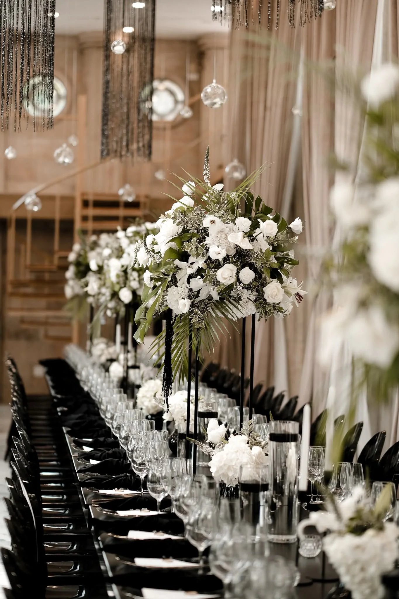 Elegant banquet table set with white and black floral centerpieces, glassware, and black chairs, decorated with hanging glass baubles in a wooden-paneled room.