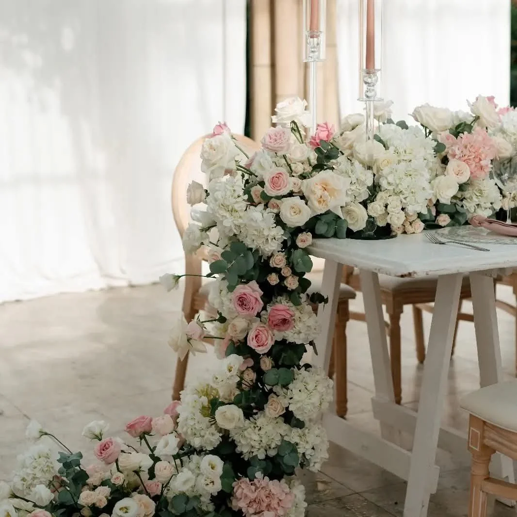 Wedding table decoration with a lush floral arrangement of white and pale pink roses, peonies, and greenery, draping over a white table, with candles and table settings.