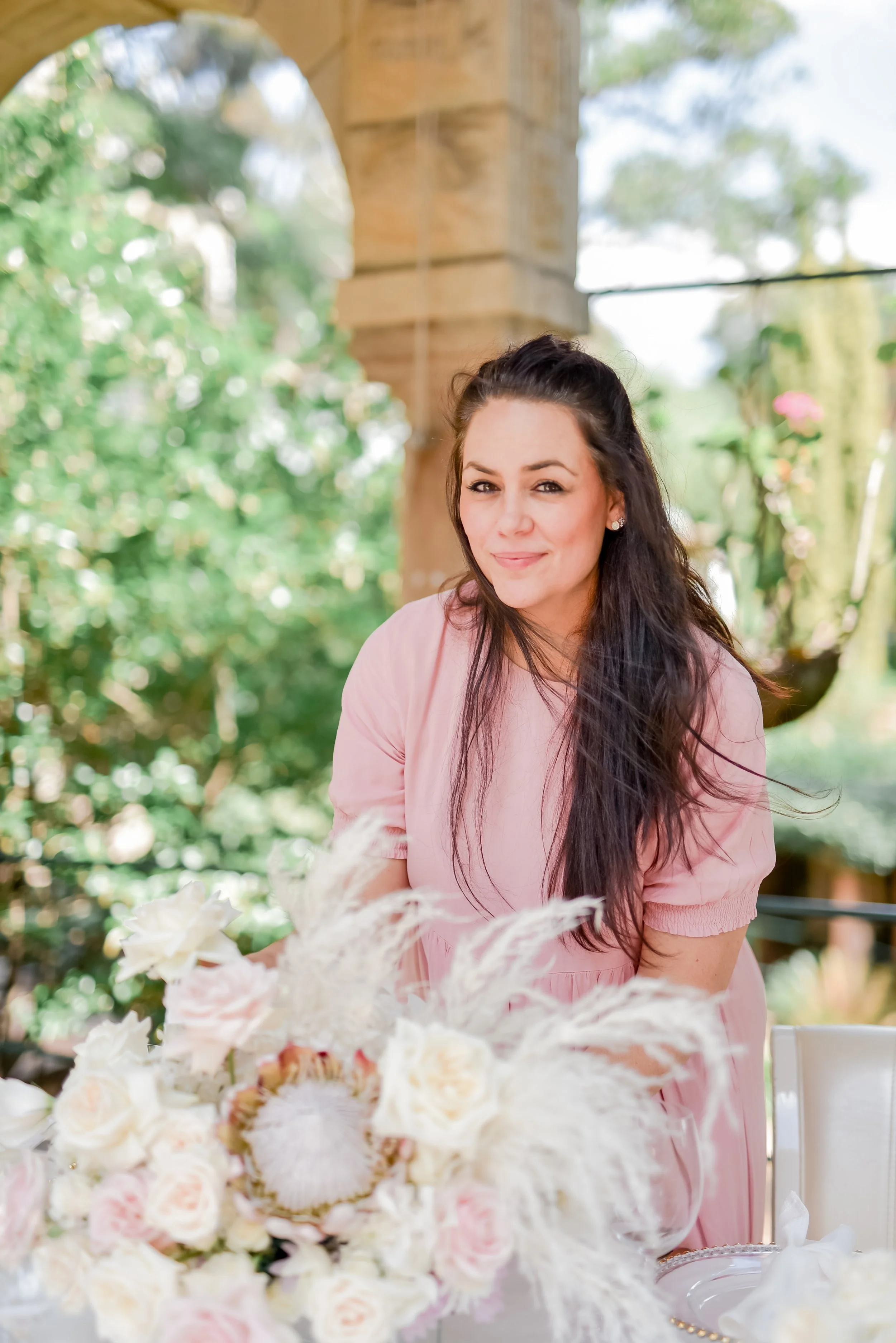 Mira Cameron in a pink dress styling at a table a large bouquet of white and pink flowers, outdoors with green trees and a stone structure in the background.