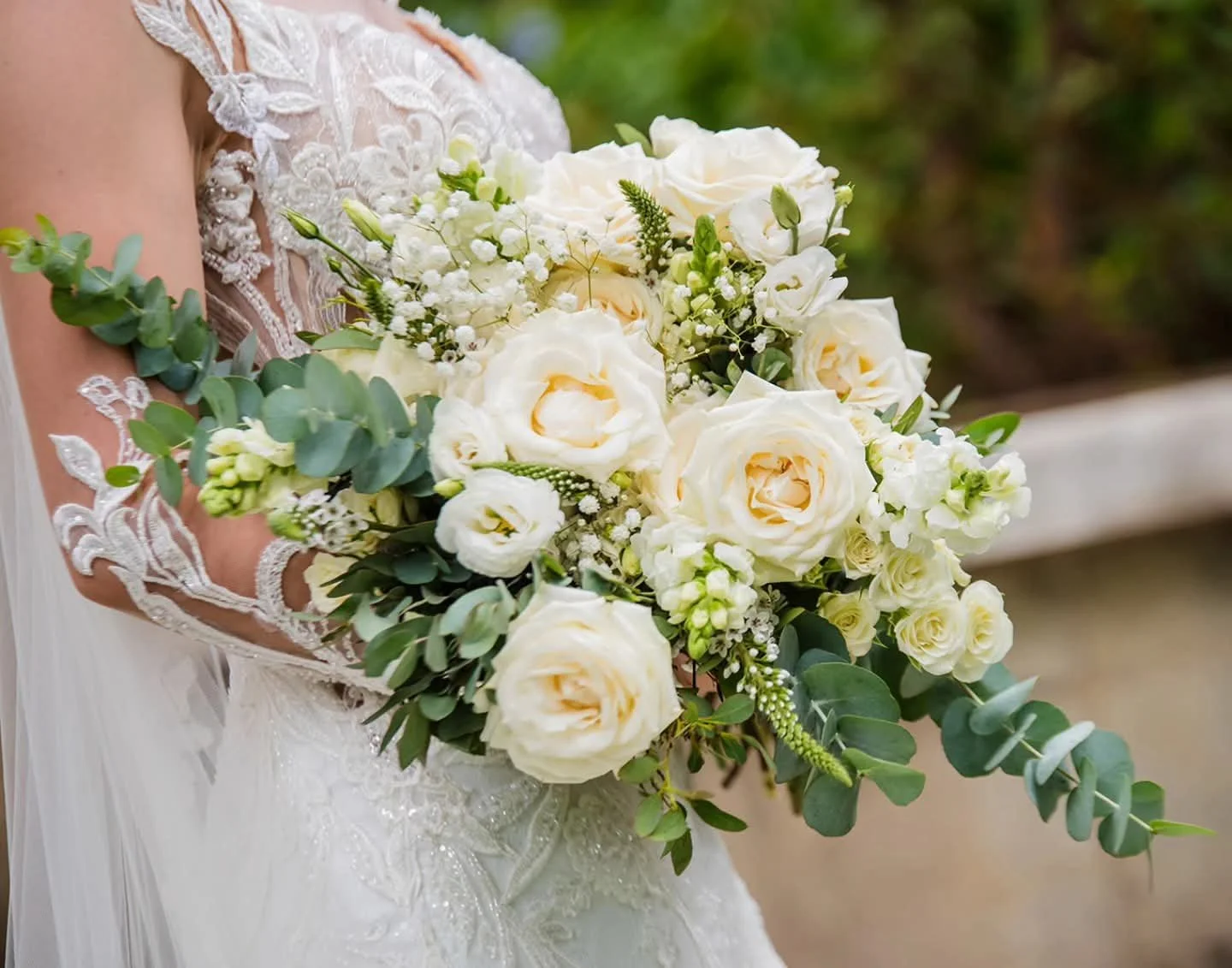 Close-up of a bride holding a bouquet of white roses, lisianthus, and eucalyptus, with lace wedding dress details visible.