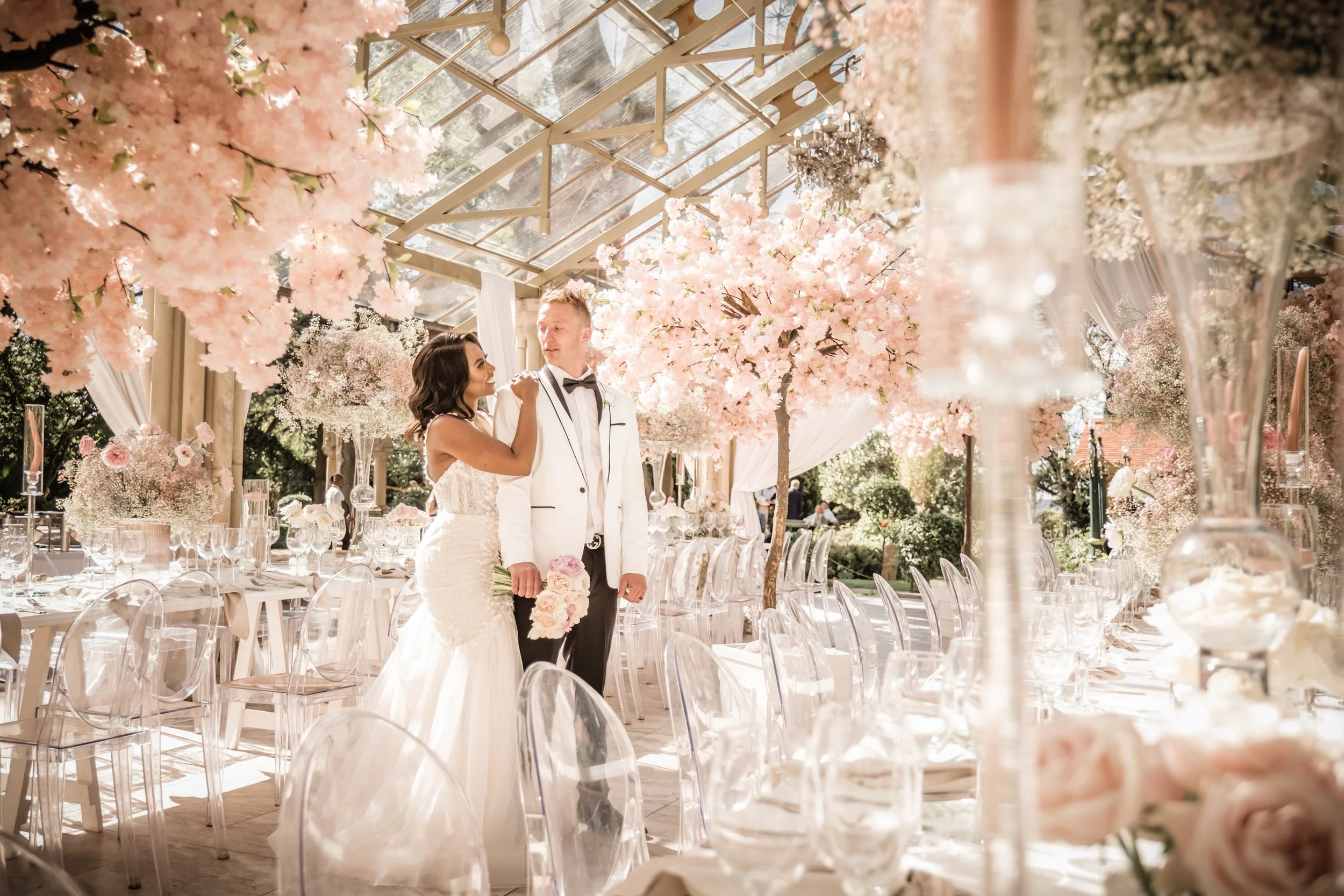 A bride and groom stand together at a wedding reception, surrounded by pink cherry blossom trees, beautiful flowers and elegant table settings, under a glass roof with sunlight streaming through.