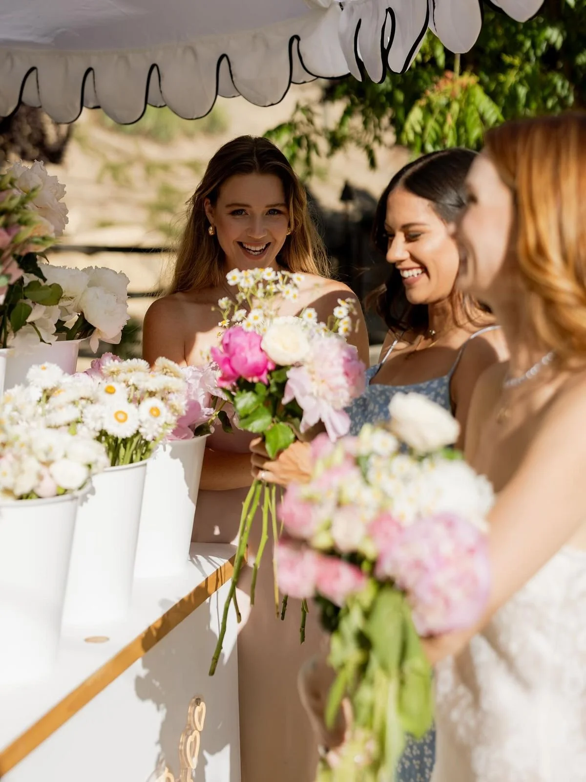 Three women smiling and looking at bouquets of pink, white, and yellow flowers at a flower bar outdoors under a white awning during a daytime event.
