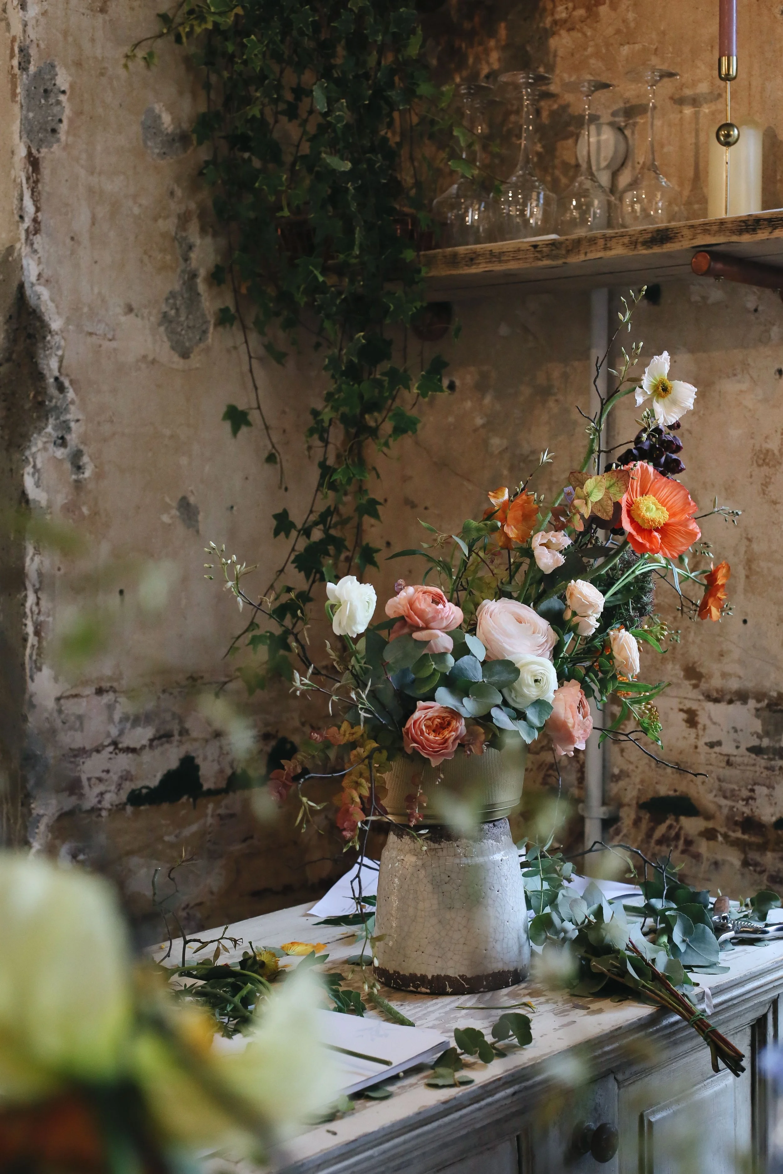 A floral arrangement with orange, white, and pink flowers and greenery in a rustic, textured ceramic vase on a wooden table against a weathered brick wall background.