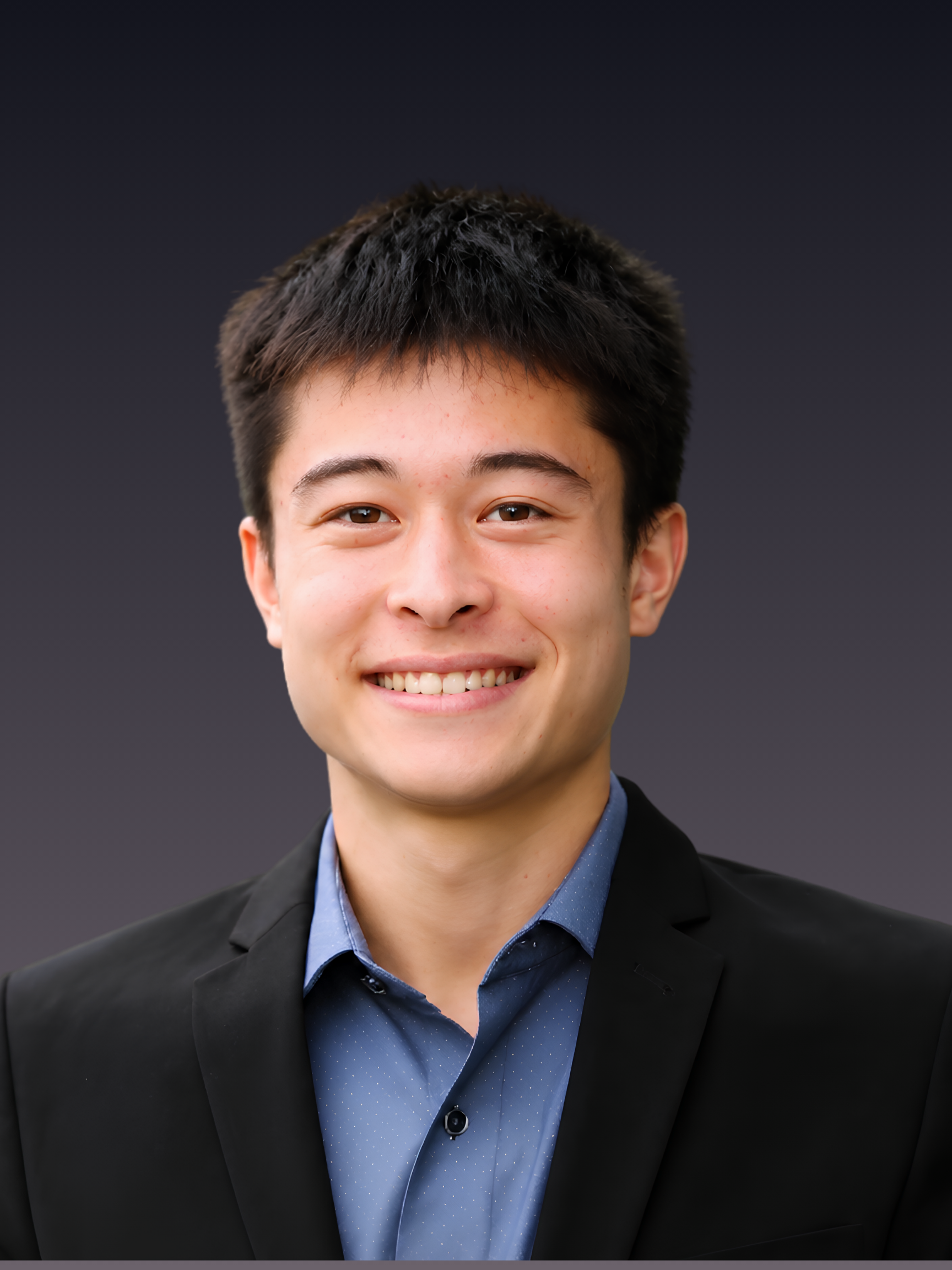 A young man wearing a black suit jacket and a blue shirt with small white dots, smiling at the camera, against a dark gray background.