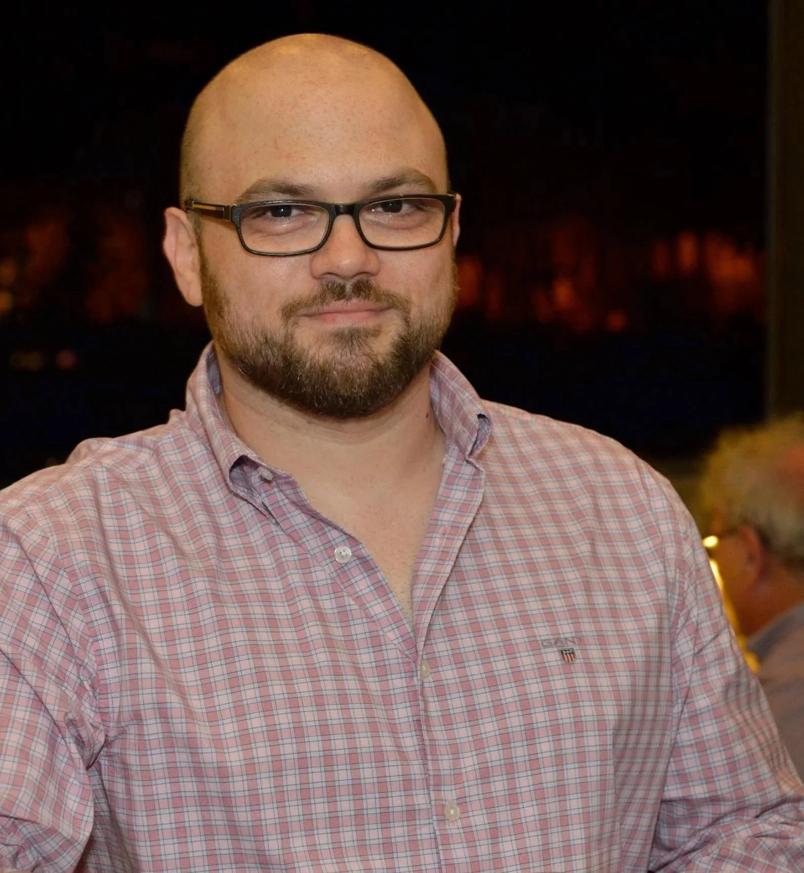 Man with glasses and beard wearing a pink checkered shirt, smiling, indoors at night.