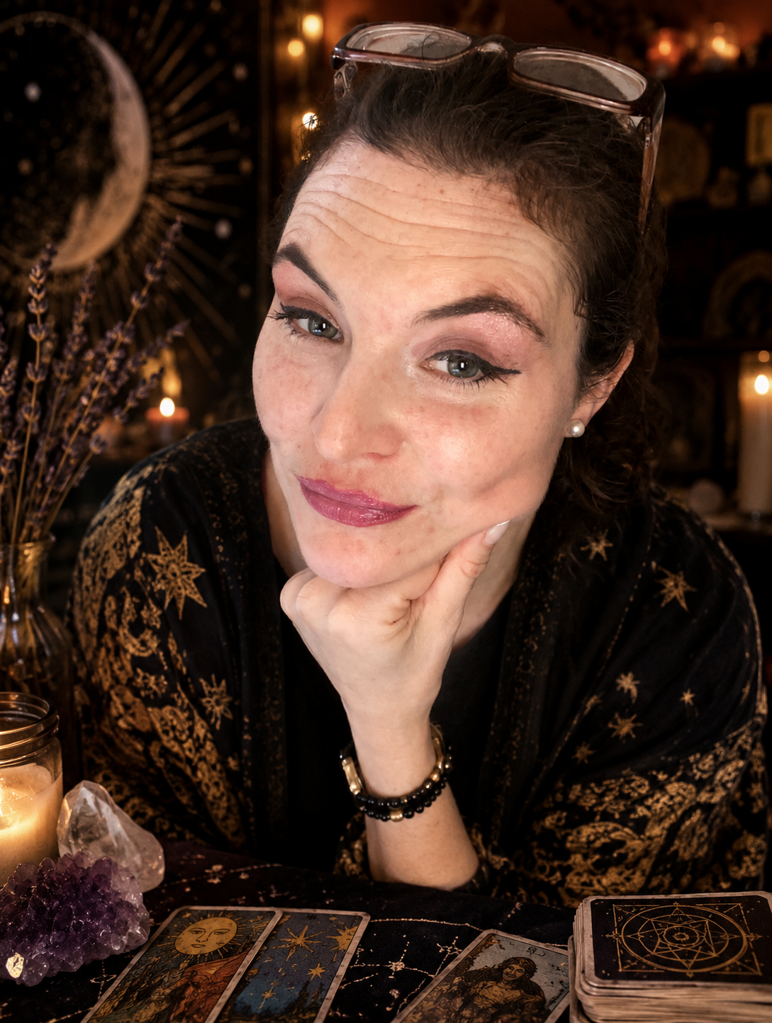 A woman with dark hair, wearing glasses on her head and pearl earrings, sits at a table with tarot cards, candles, and crystals, in a dimly lit, mystical setting.