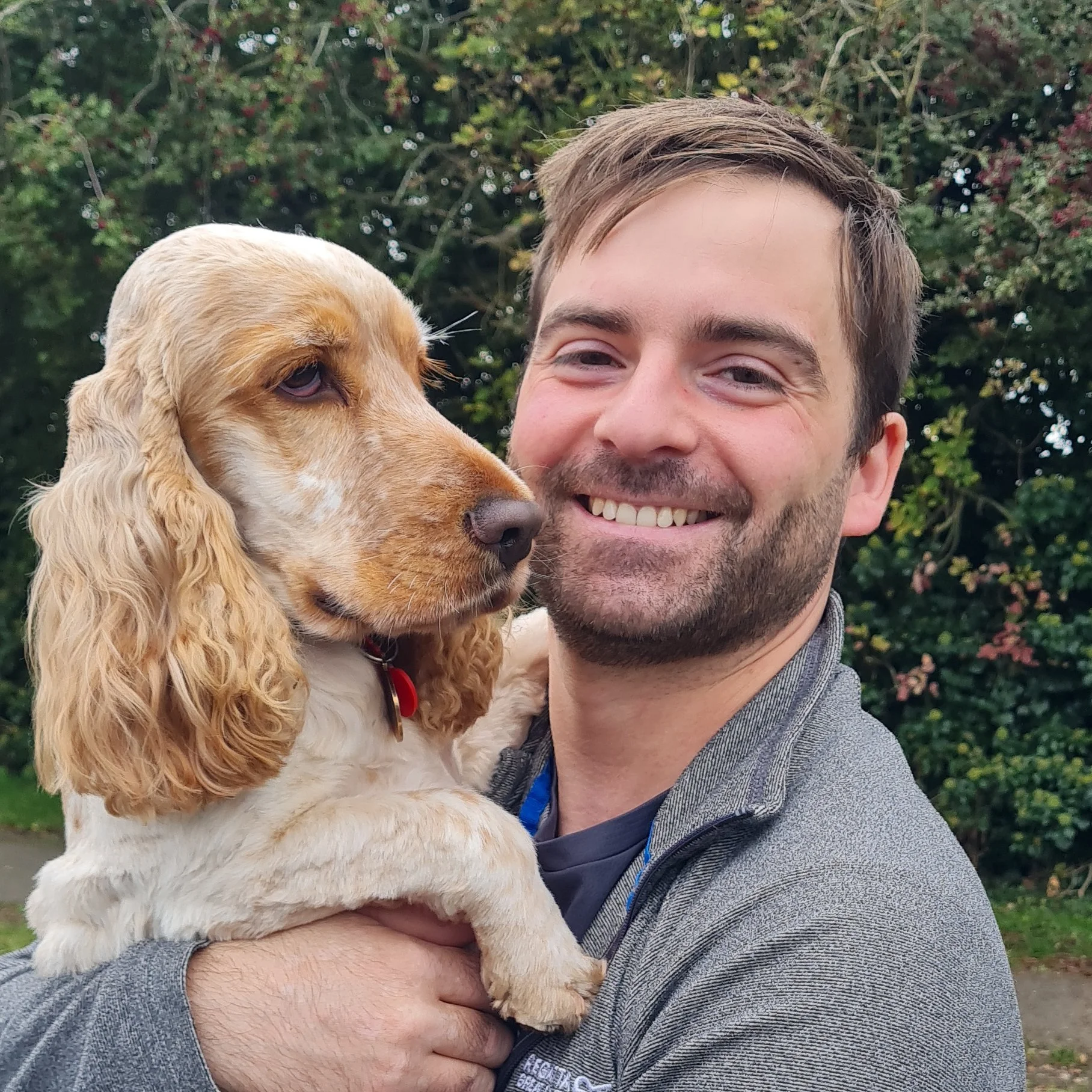 A man with short brown hair and a beard holding a light brown and white cocker spaniel dog in front of green foliage.