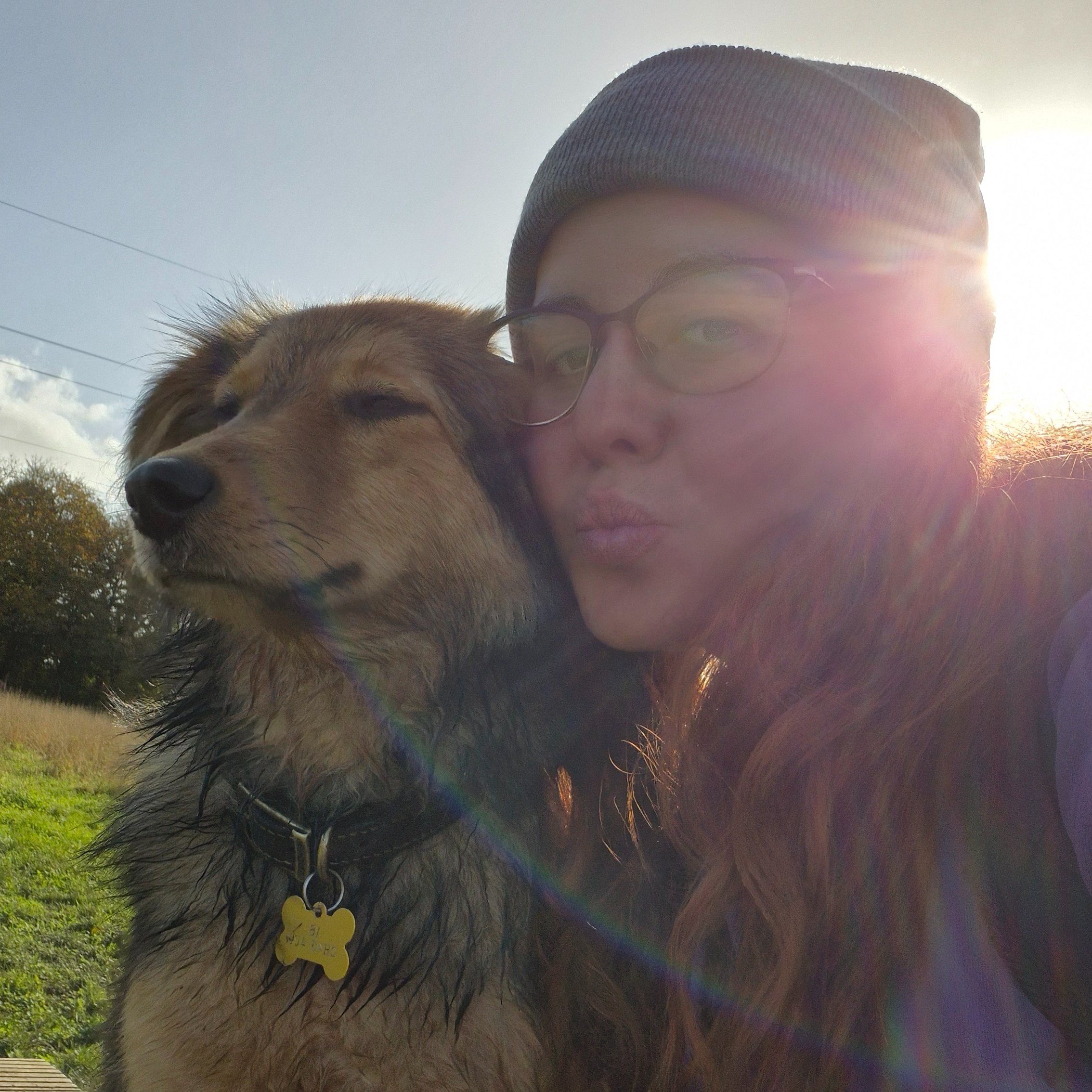 A person wearing a gray beanie and glasses taking a selfie with a dog outdoors during sunset, with trees and power lines in the background.