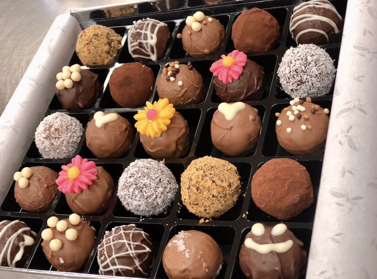 Schachtel mit verschiedenen handgefertigten Schokoladen-Trüffeln und Pralinen, einige dekoriert mit Blumen, weißen Schokoladen-Detail und Kokosraspeln.