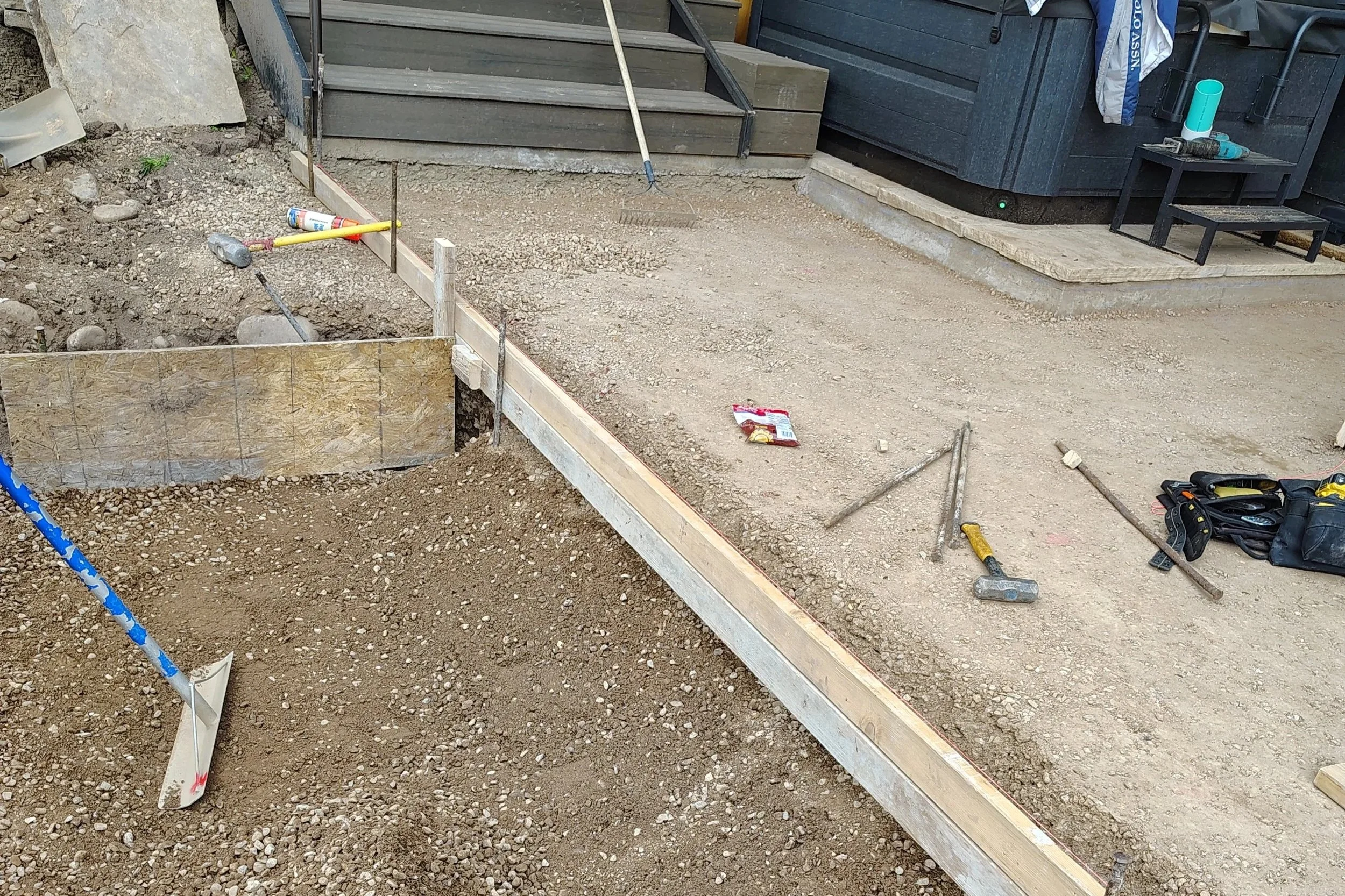 A construction site with dirt and gravel, wooden framing for a pathway or border, and various tools scattered around, including a hammer, drill, and level, near a set of stairs and a small black bench with items on top.