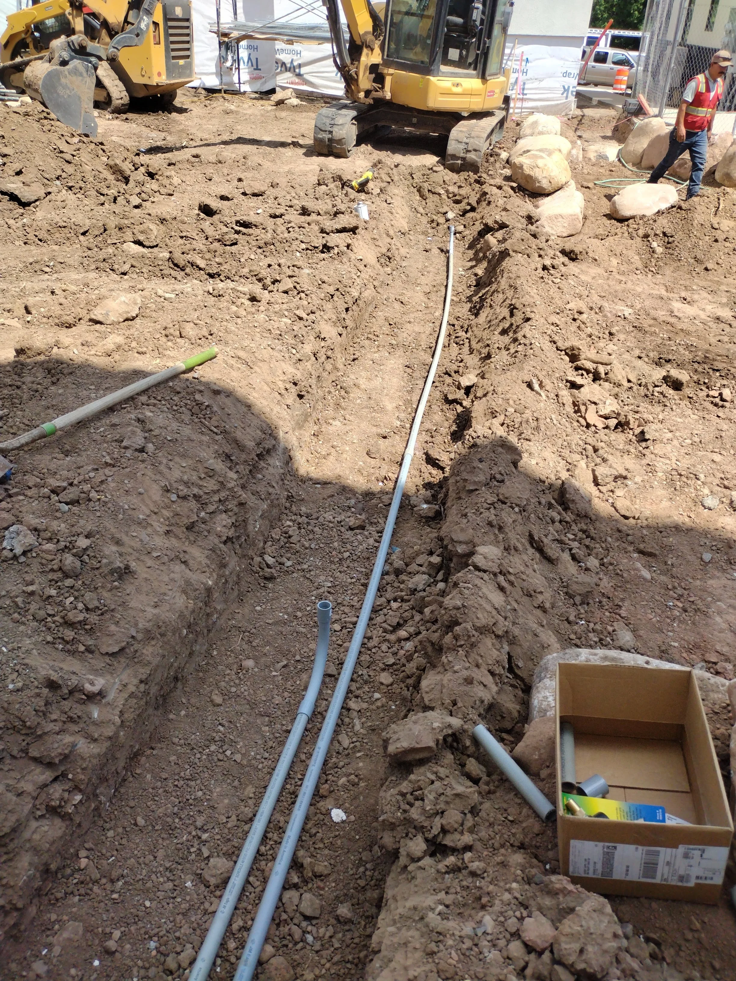 Construction site with excavator and construction worker, trenches with pipes, rocks, and construction supplies.
