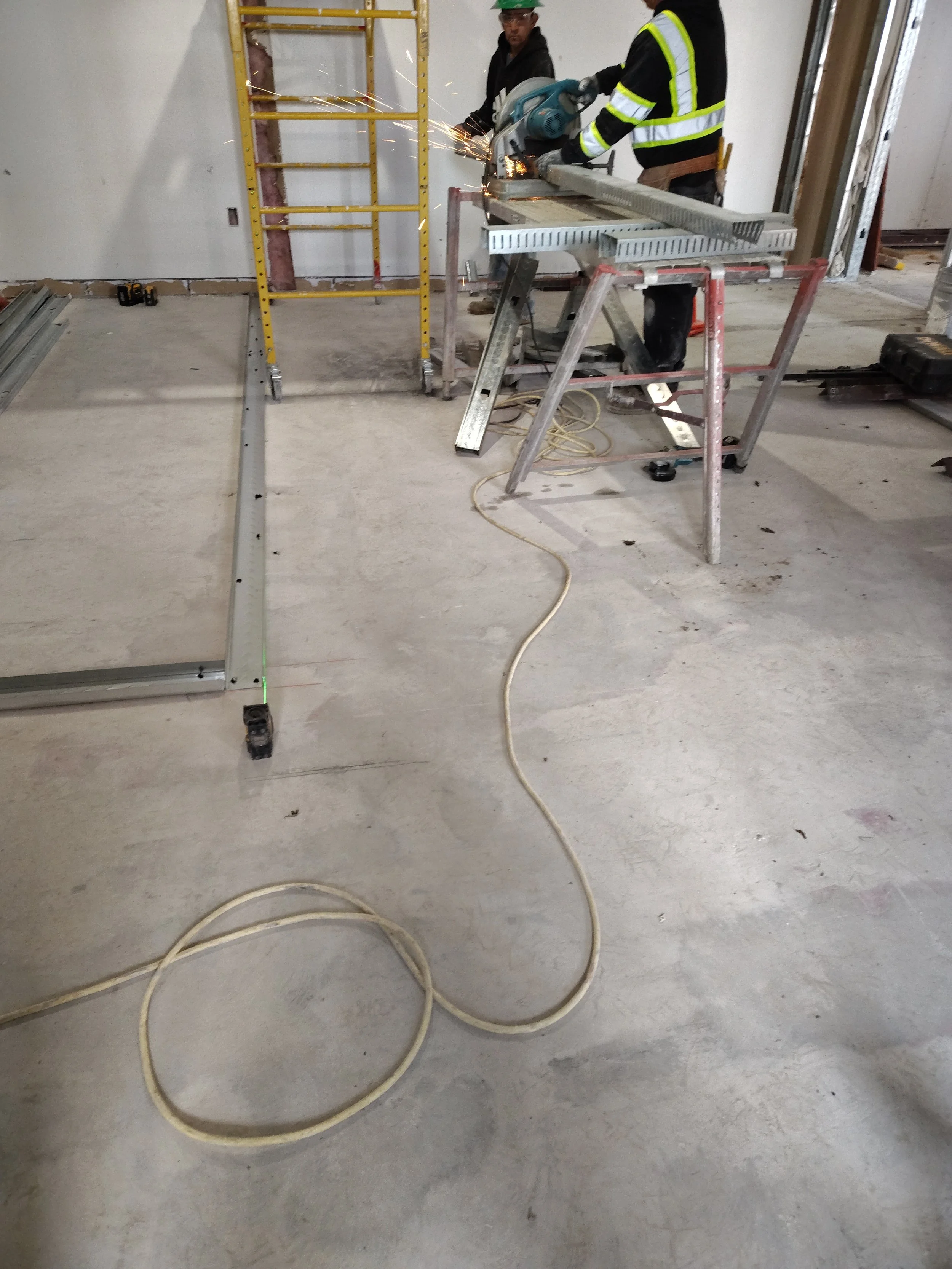 Two construction workers, one in a high-visibility safety jacket and the other in dark clothing, are using a circular saw to cut a piece of material in an unfinished room. Sparks are flying from the saw, and there is construction equipment and tools scattered around, including a ladder, a long piece of metal on the floor, and power cords.