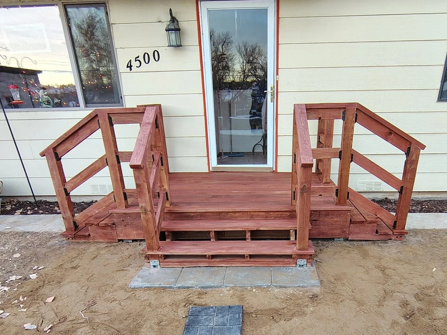 Newly built wooden ramp with handrails outside a house, leading to a door, with steps at the entrance, set on a dirt yard.