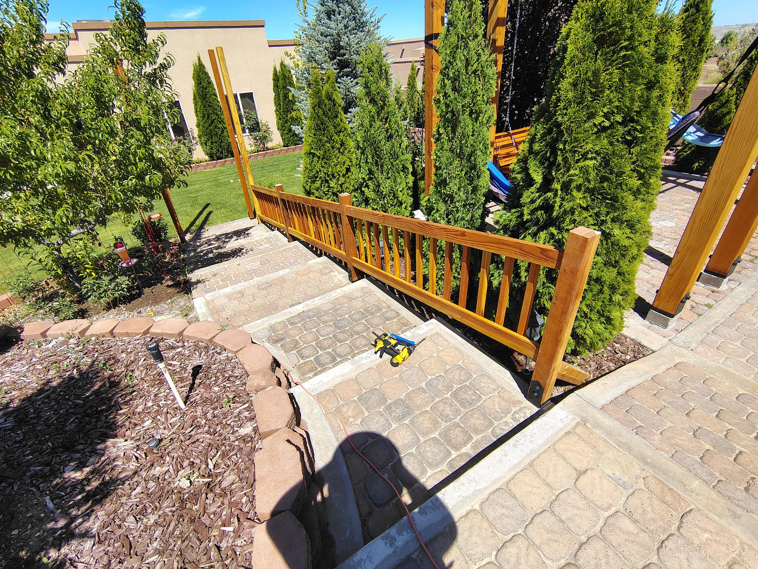 Backyard garden with stone steps, new wooden fence, tall green trees and shrubs, and tools on the ground.