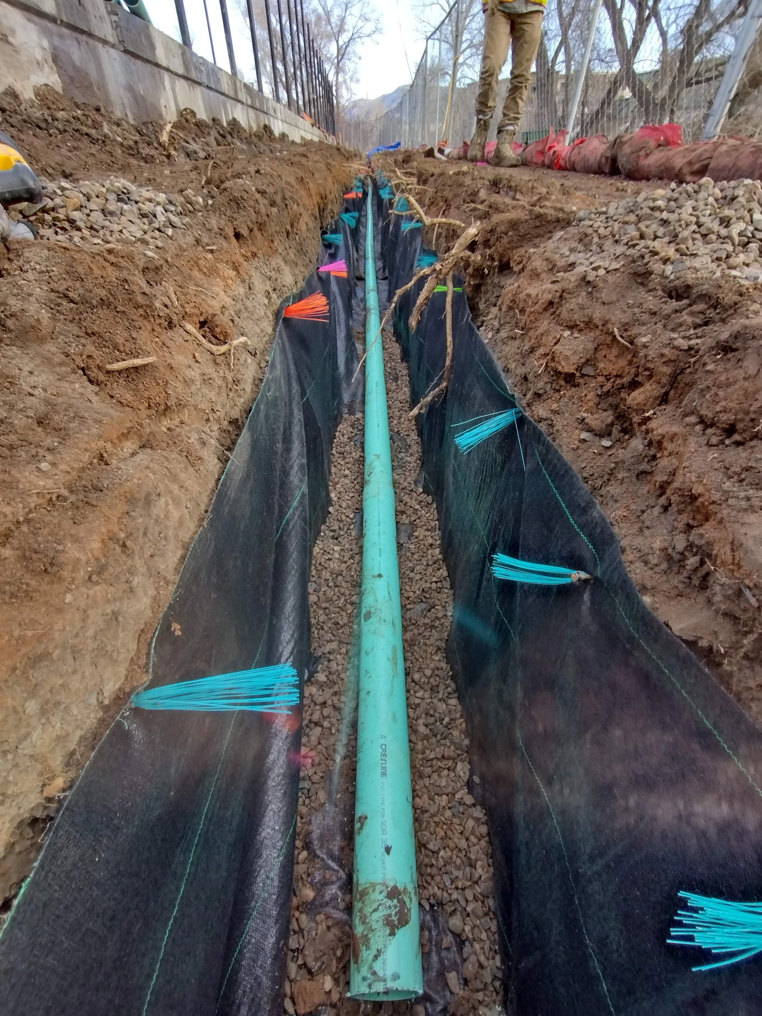A trench with a green pipe in the center, reinforced with black fabric and colorful zip ties, with a worker in the background and fencing on both sides.