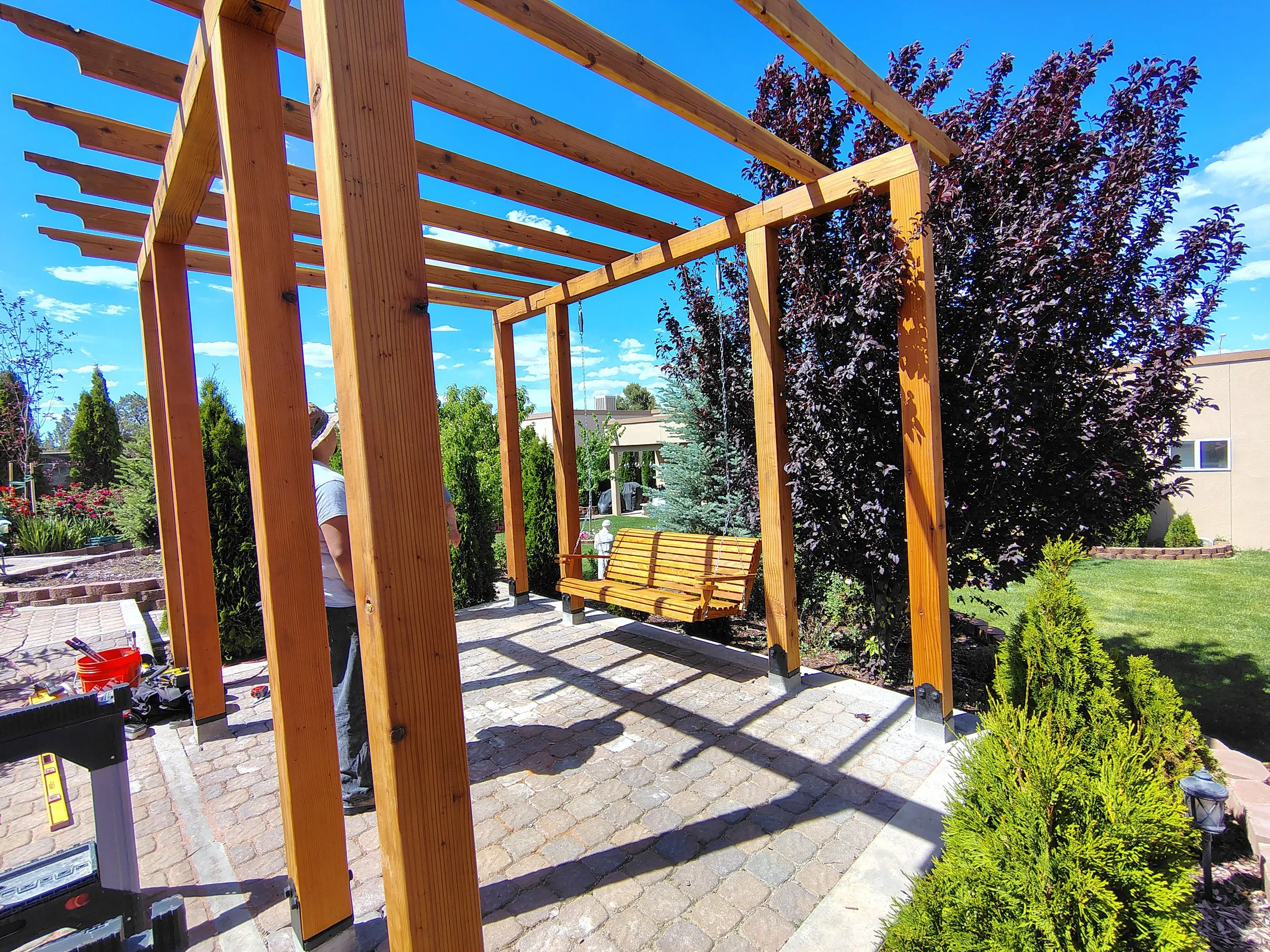 Wooden pergola under construction in a backyard garden with a yellow bench, lush trees, and a blue sky.